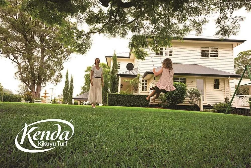 A woman and child enjoying a backyard swing set in front of a large white house and green lawn. Kenda Kikuyu Turf logo.
