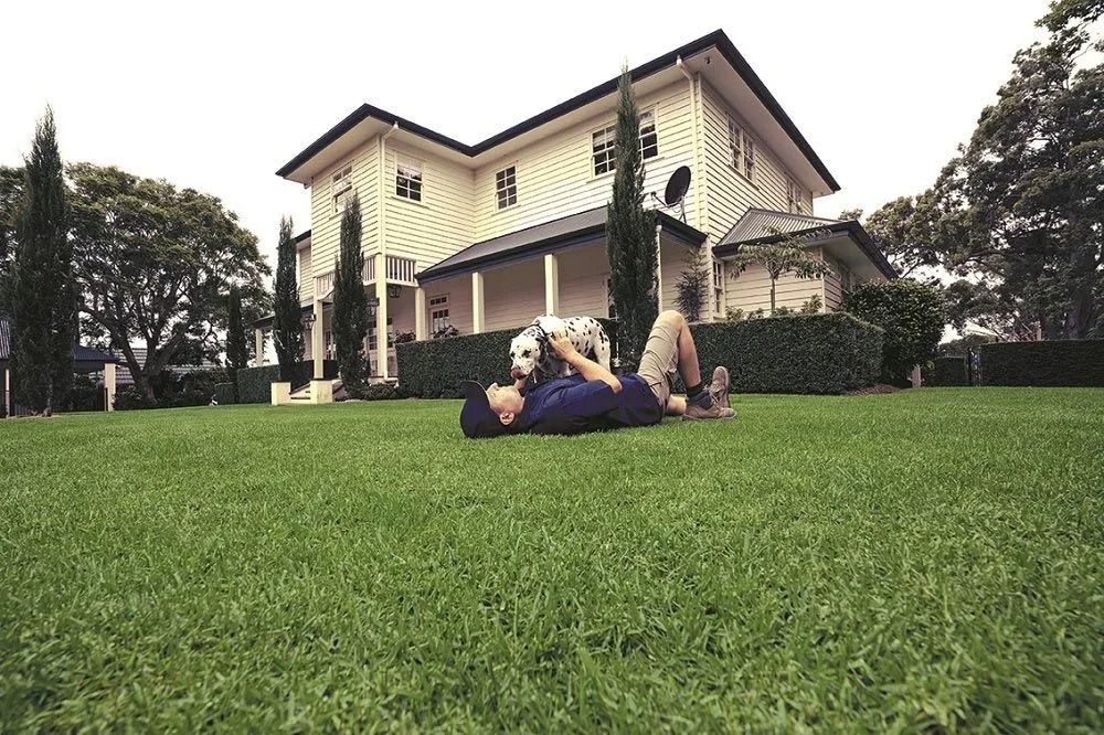Man lying on a green lawn, petting a dog, in front of a two-story house with a white exterior. Overcast sky.