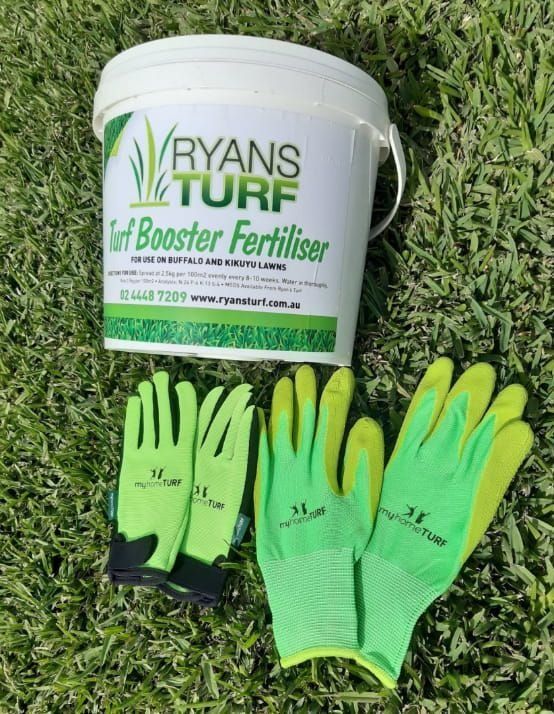 A bucket of Ryans Turf Booster Fertiliser sits on grass with two pairs of green garden gloves.