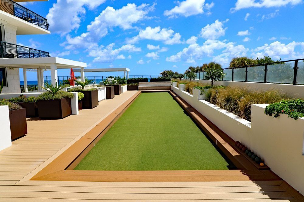 Outdoor bocce ball court with green turf, surrounded by wood decking and white walls, under a blue sky.