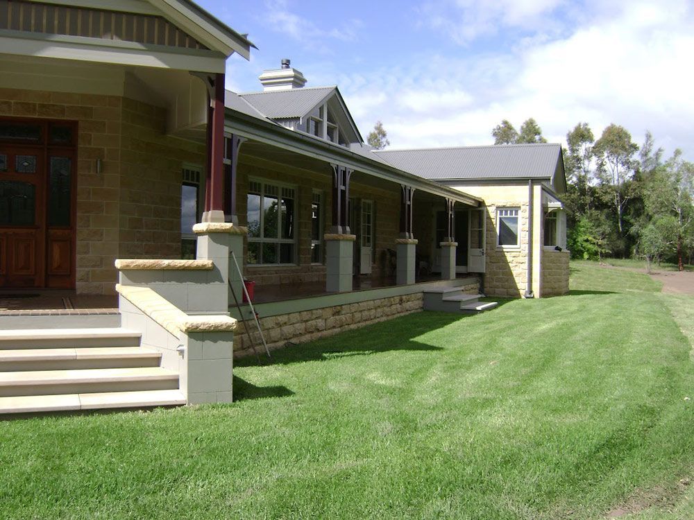 A beige stone house with a covered porch, steps, and a green lawn on a sunny day.