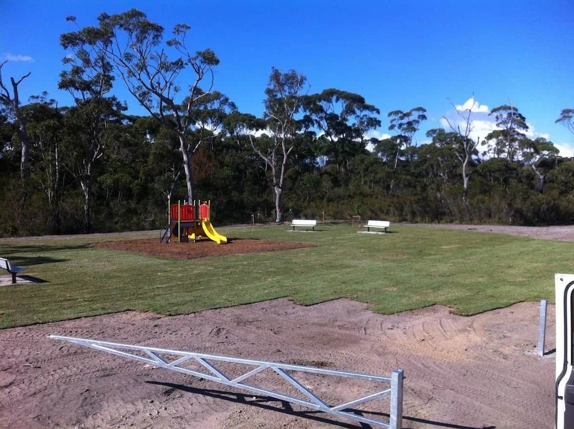 A children's playground with a slide and swing set on a grassy area, surrounded by trees, under a bright blue sky.
