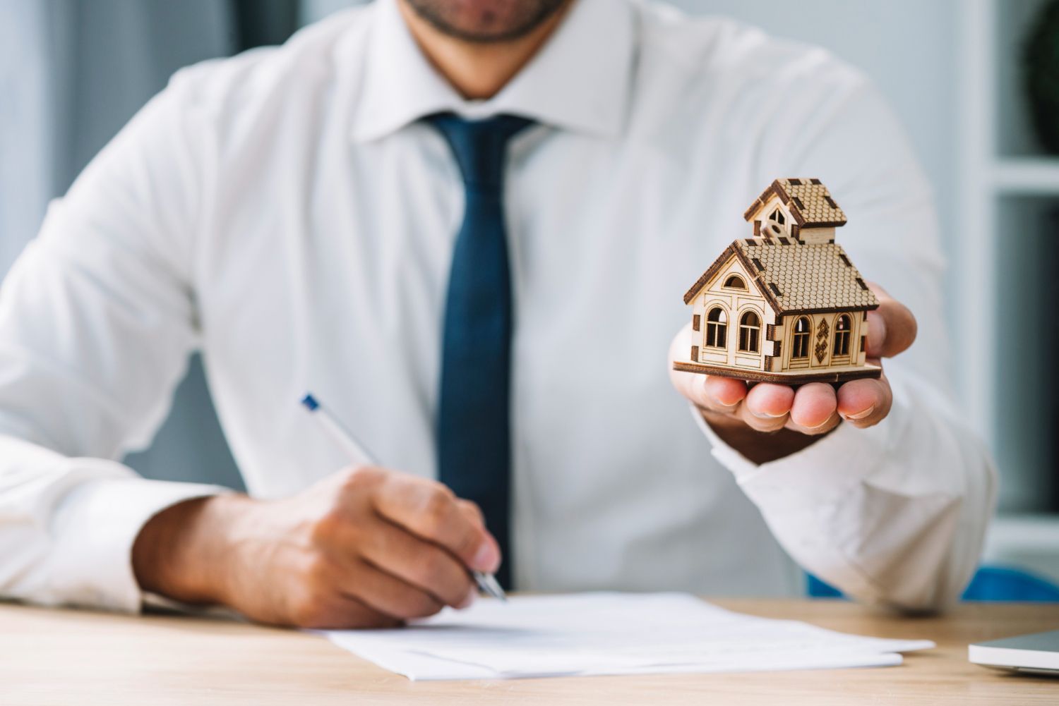 A man is holding a small wooden house in his hand while signing a document.