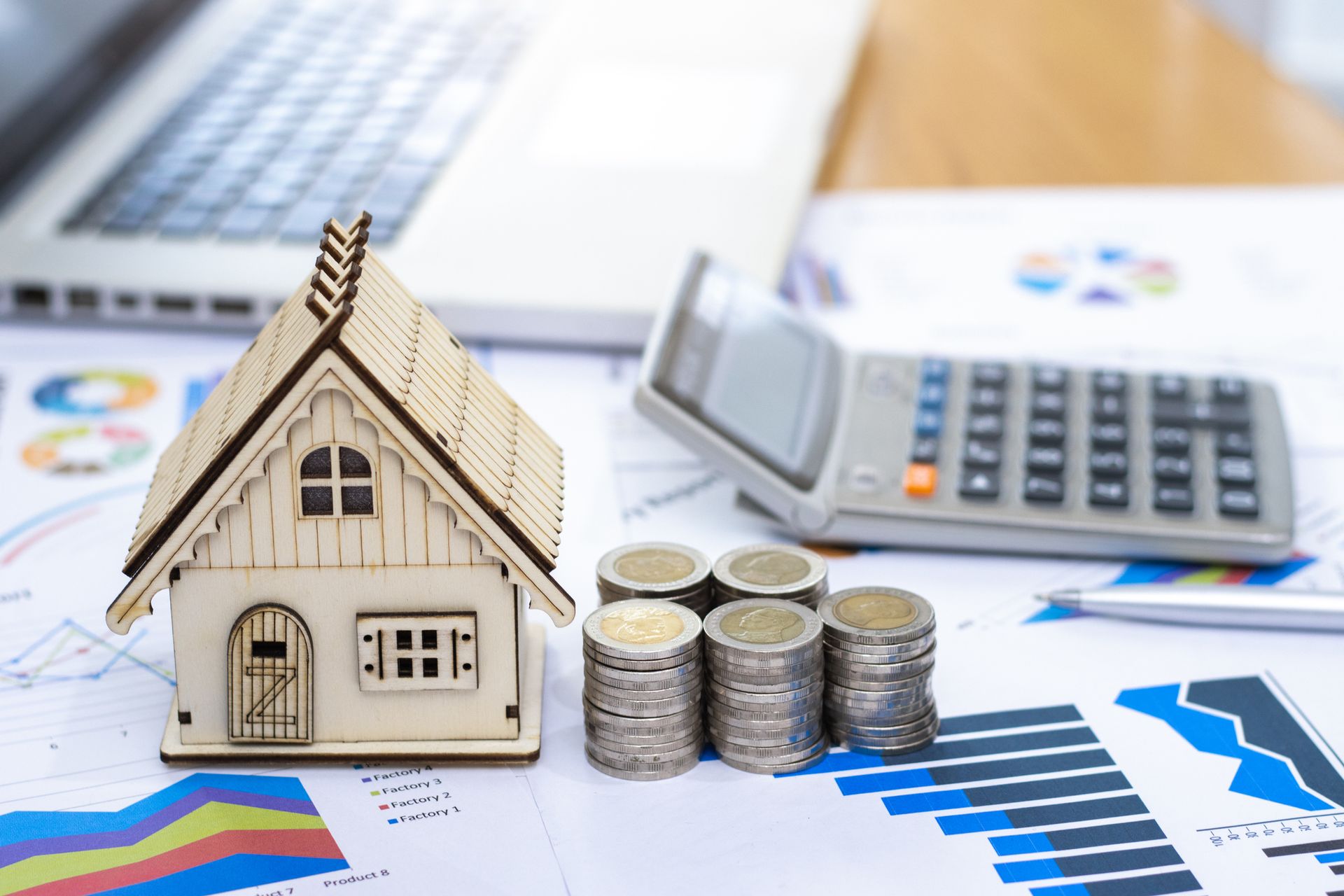 A wooden house is sitting on top of a table next to stacks of coins and a calculator.