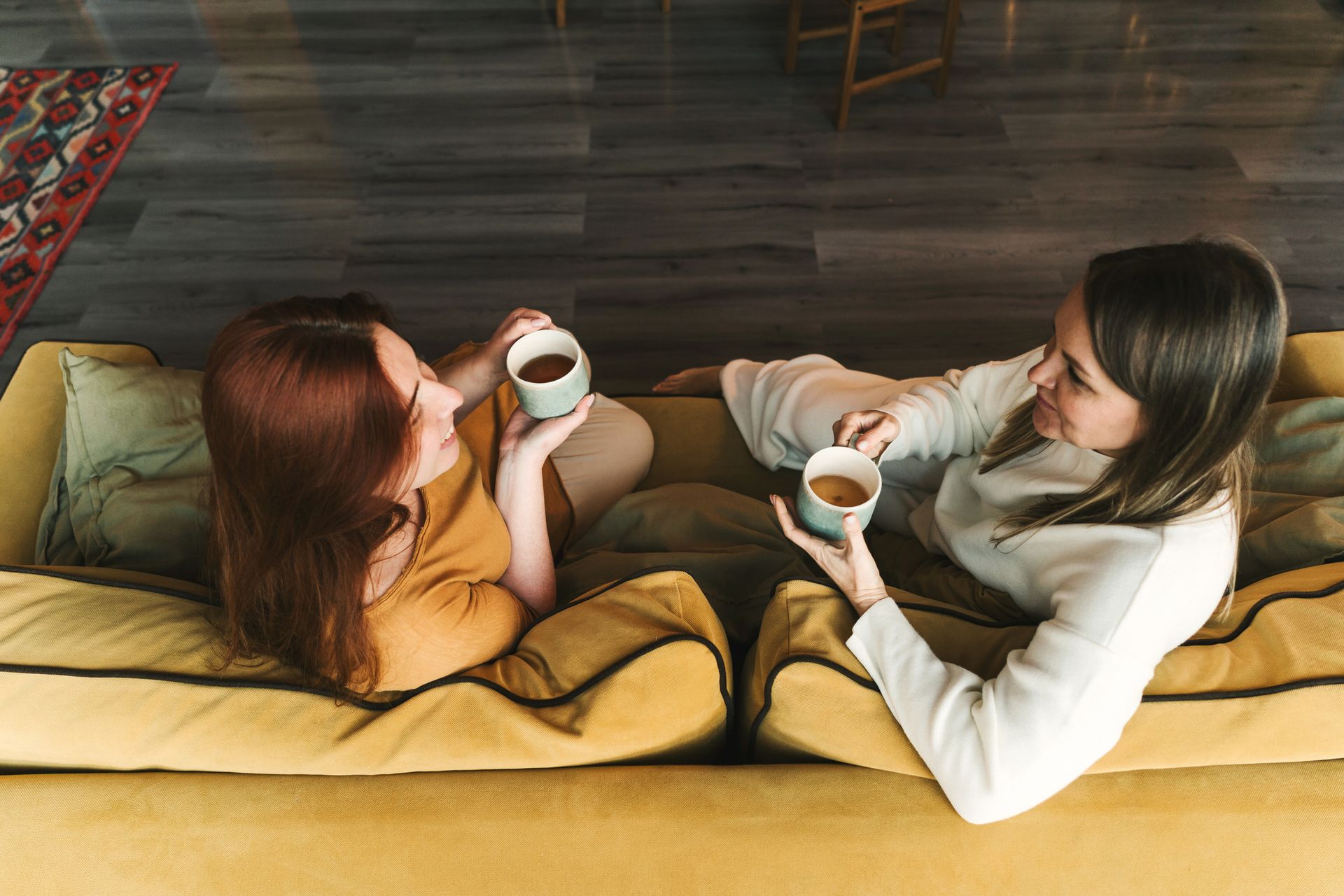 Two women on a yellow couch, holding mugs and smiling at each other. Coffee cups, indoor setting.