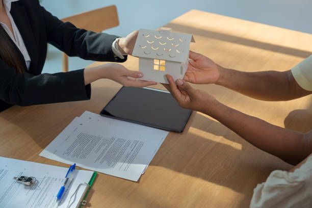 Person handing a model house to another person, over a table with documents.