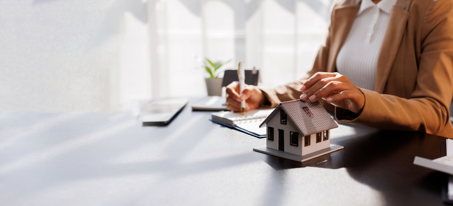 A person in a blazer touching a miniature house on a desk while writing.