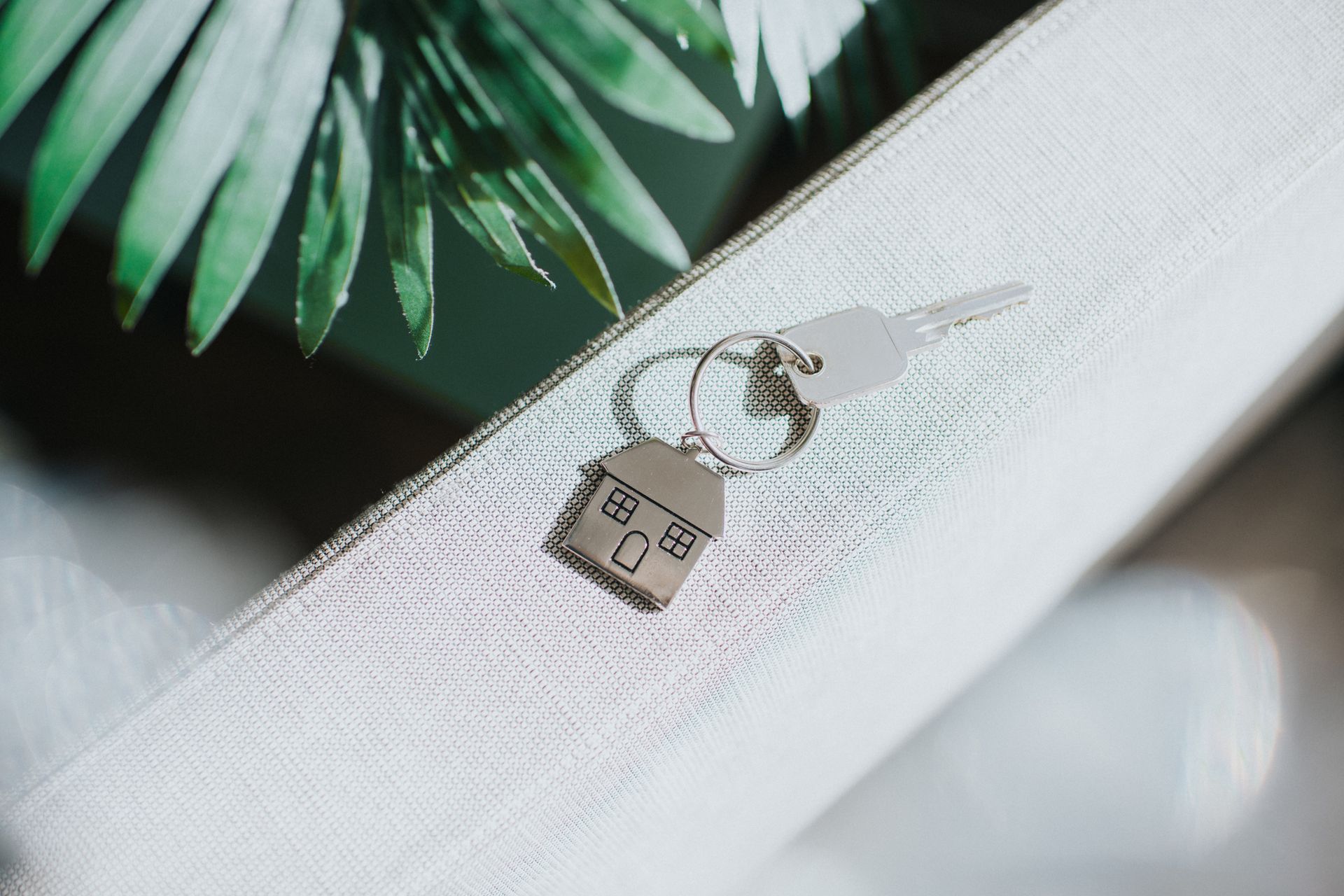 Key with house-shaped charm resting on white fabric, with a leafy green plant in the background.