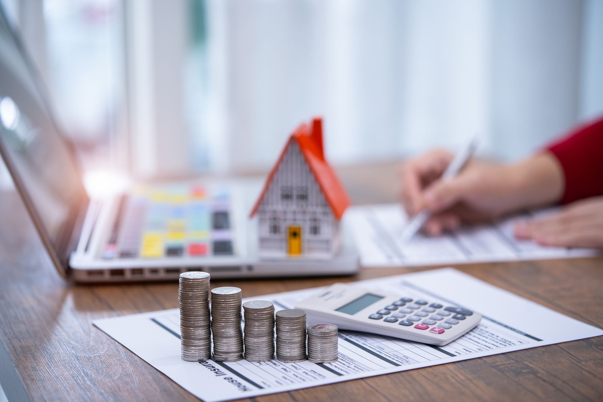 Stacks of coins, a toy house, calculator, and person writing; all on a table, suggesting home finances.