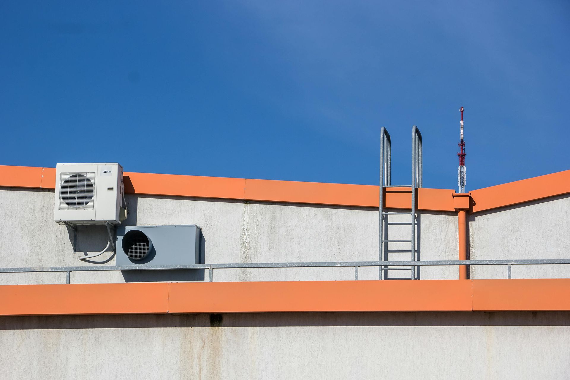 Air conditioning units, ladder, and antenna on a white building roof with orange trim against a clear blue sky.