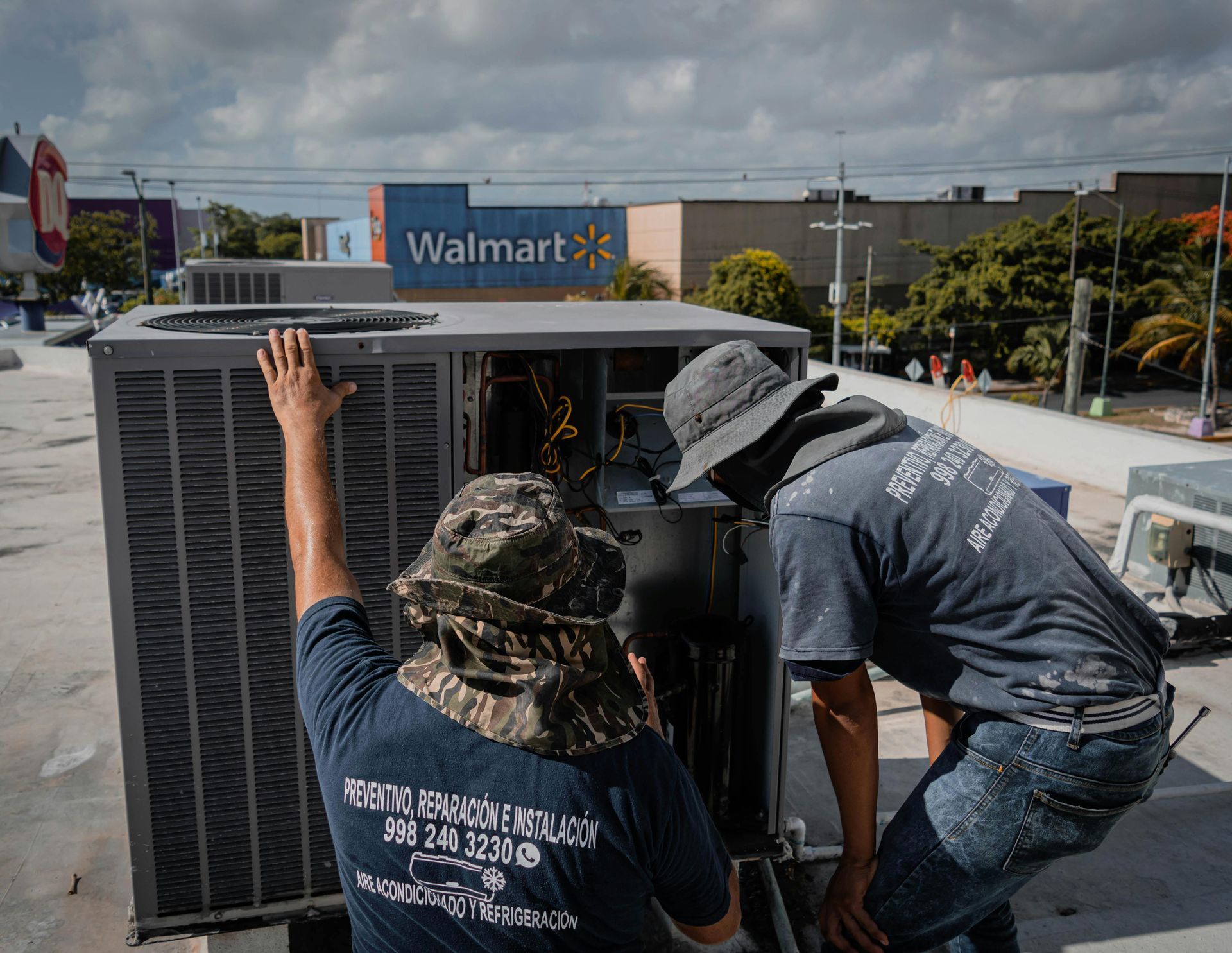 Two men in hats repair an AC unit on a roof with a Walmart in the background on a sunny day.