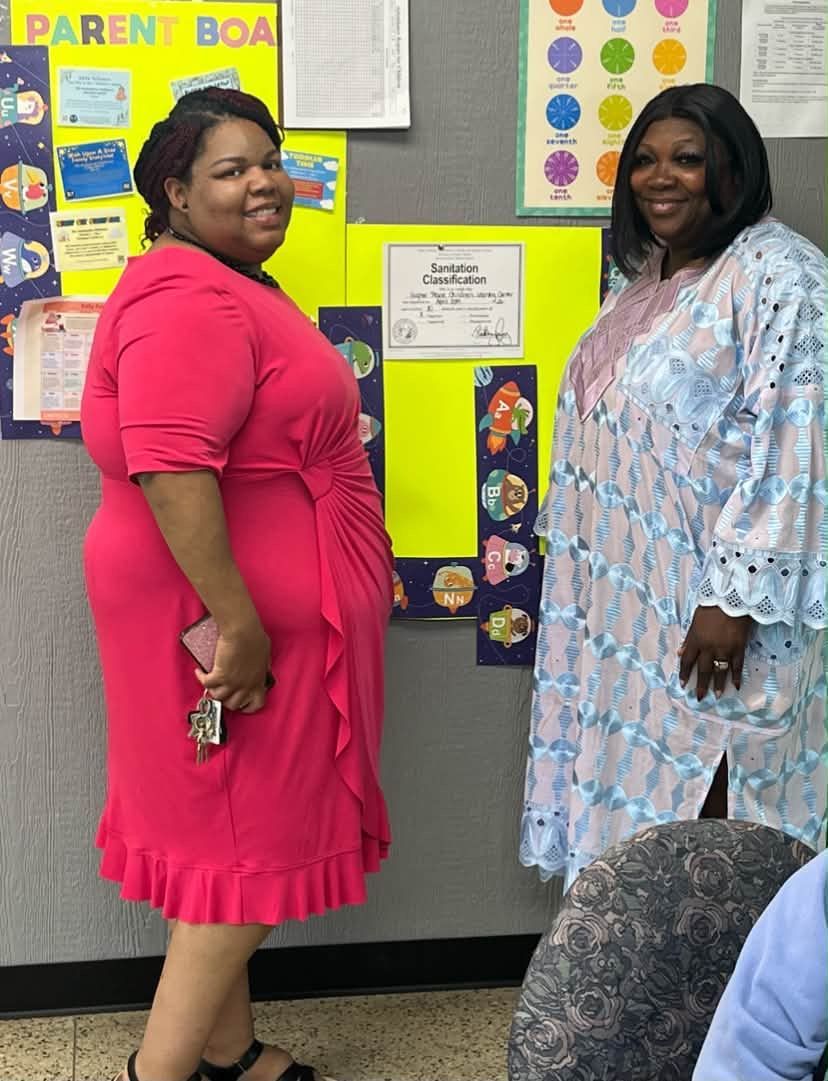 Two women smiling in front of a bulletin board covered with colorful classroom posters.