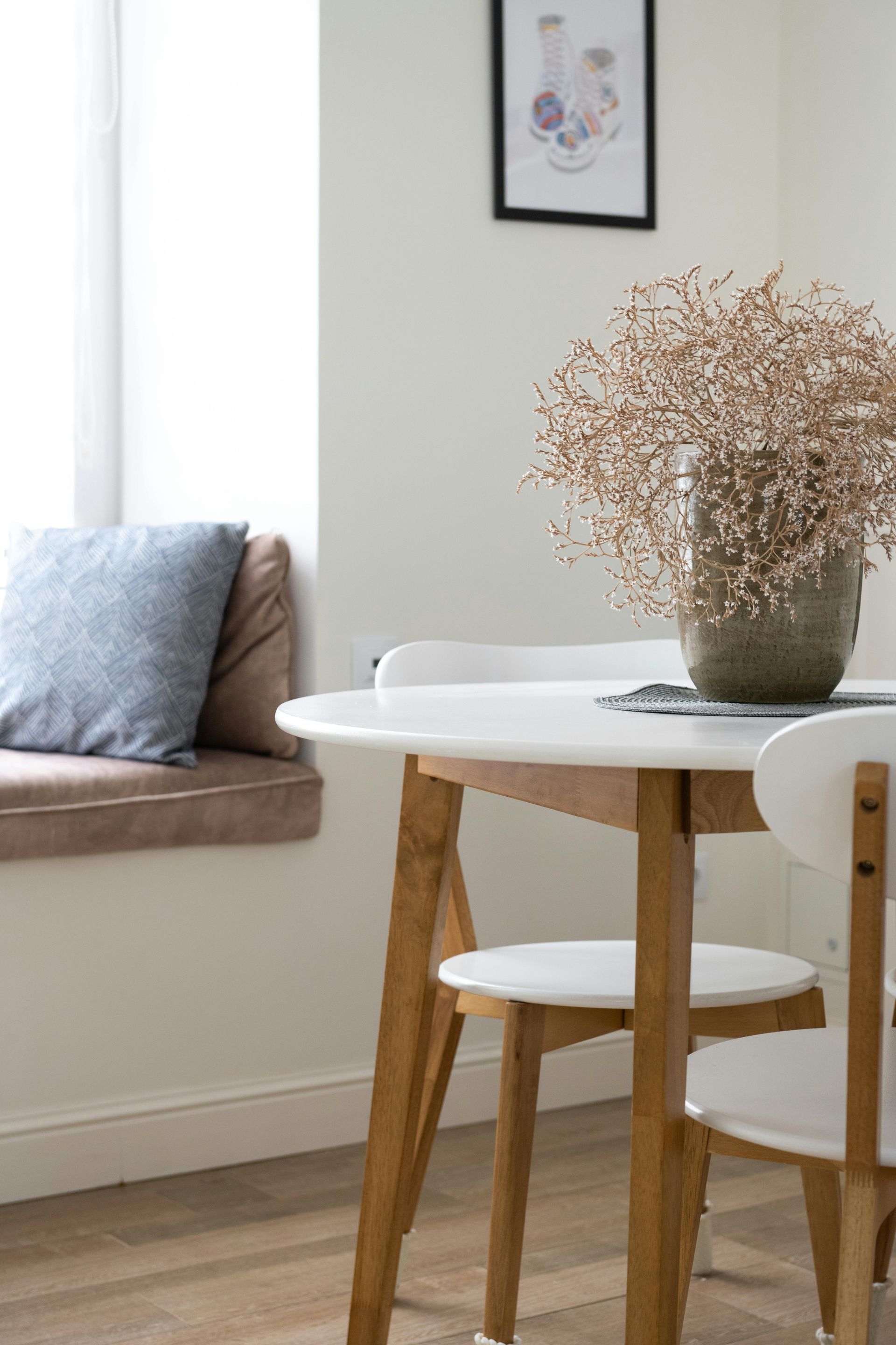 A dining room table and chairs with a vase of dried flowers on it.