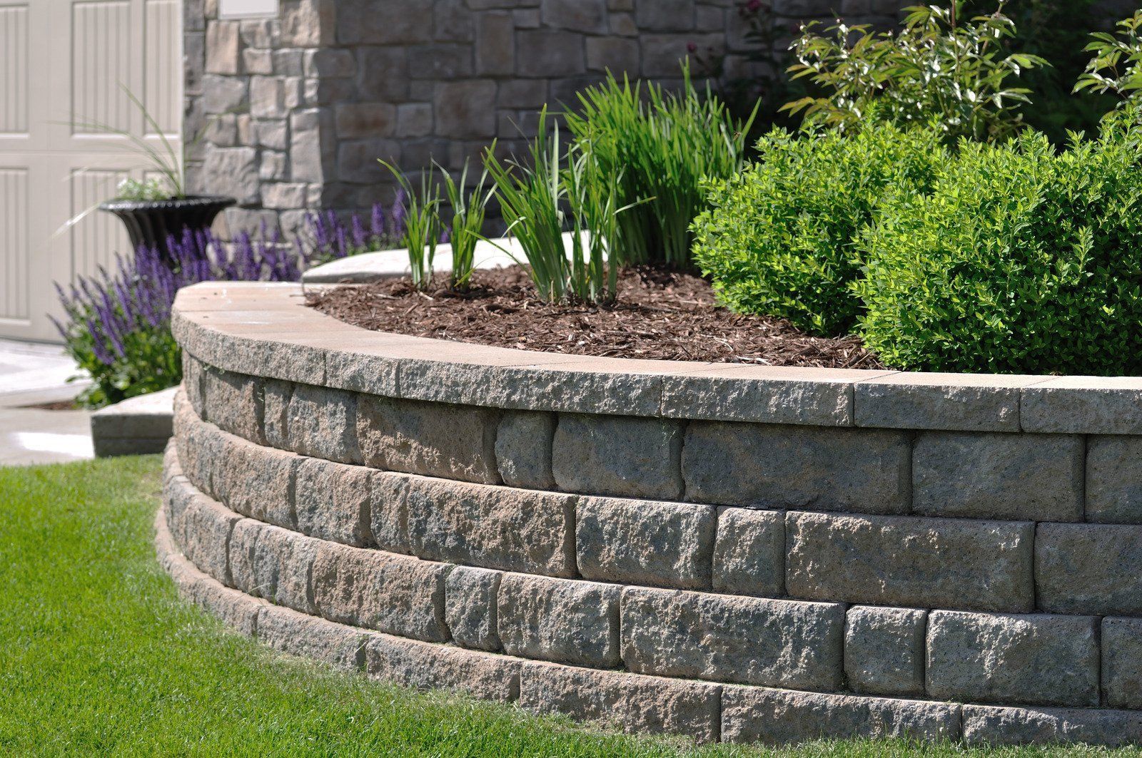 Curved stone retaining wall with a garden bed featuring plants and mulch, beside green grass.
