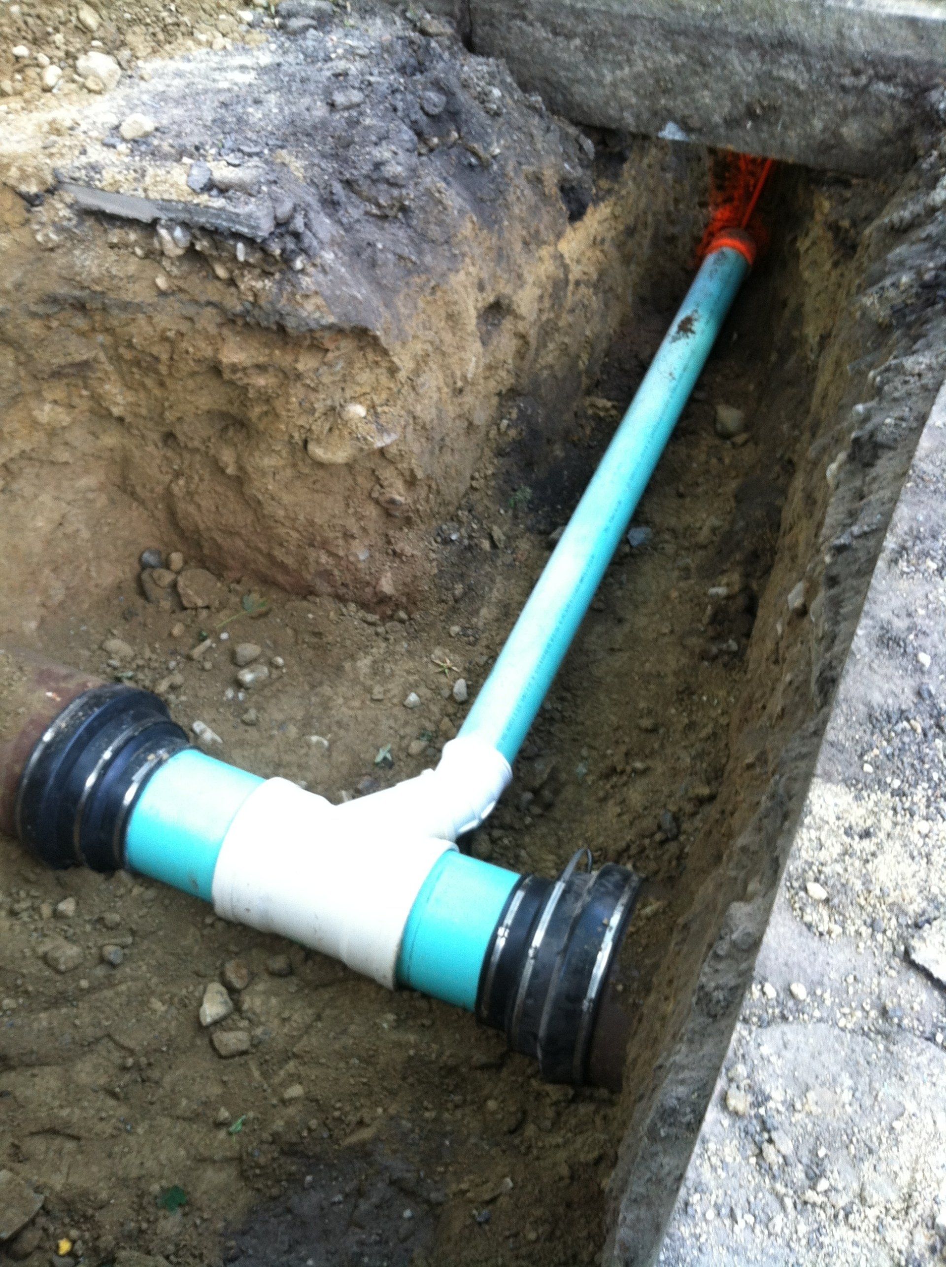 An open trench reveals a blue PVC pipe with a Y-connection, leading to a red pipe.