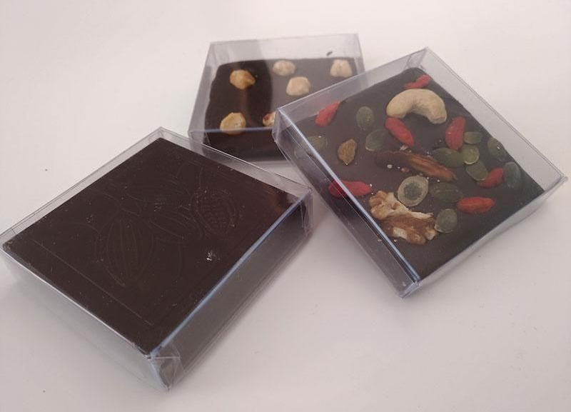 Tre barrette di cioccolato quadrate in scatole trasparenti, una semplice, le altre ricoperte di noci e semi.