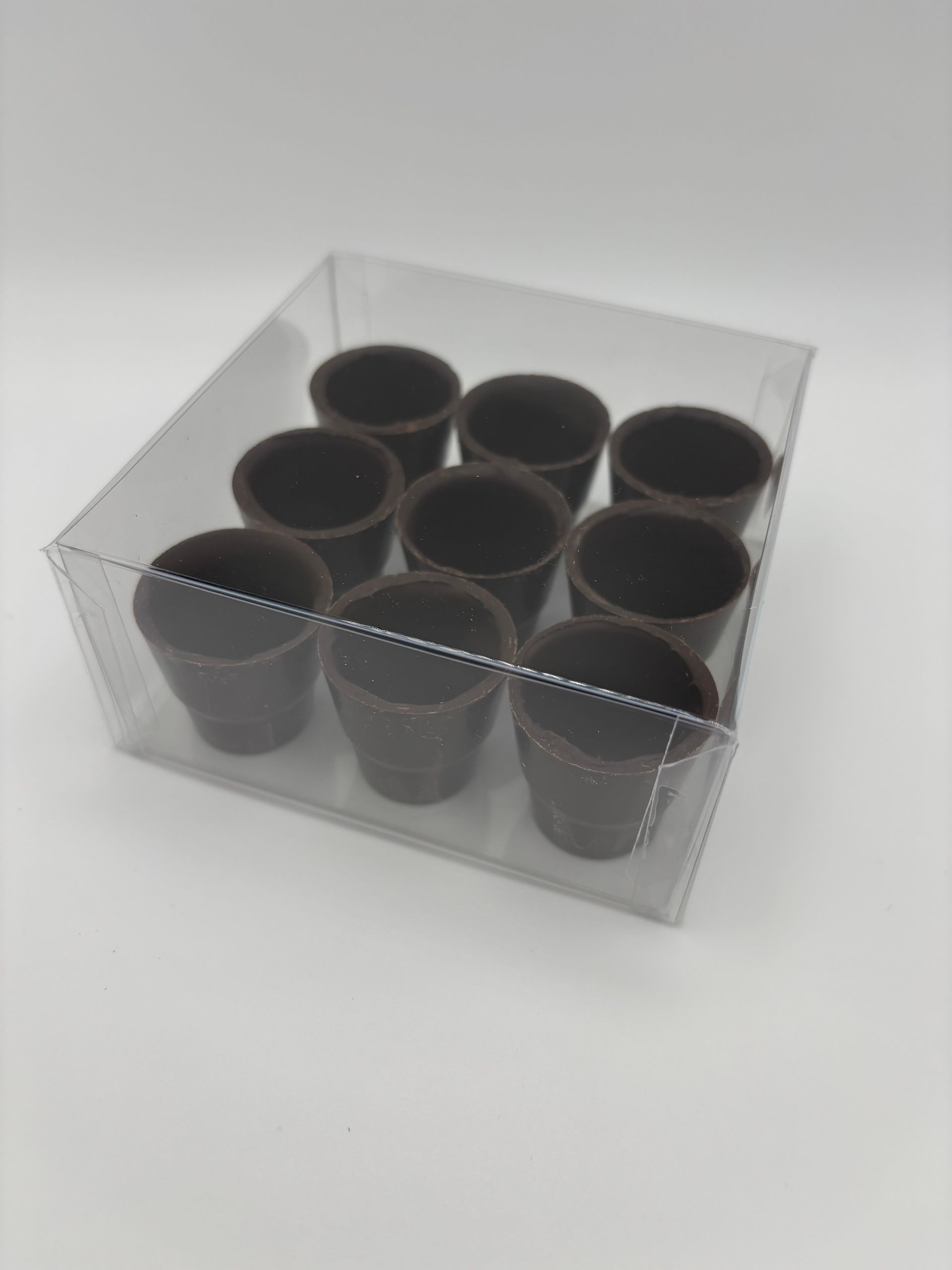 Nove tazzine di cioccolato fondente in una scatola di plastica trasparente, disposte in file su una superficie bianca.