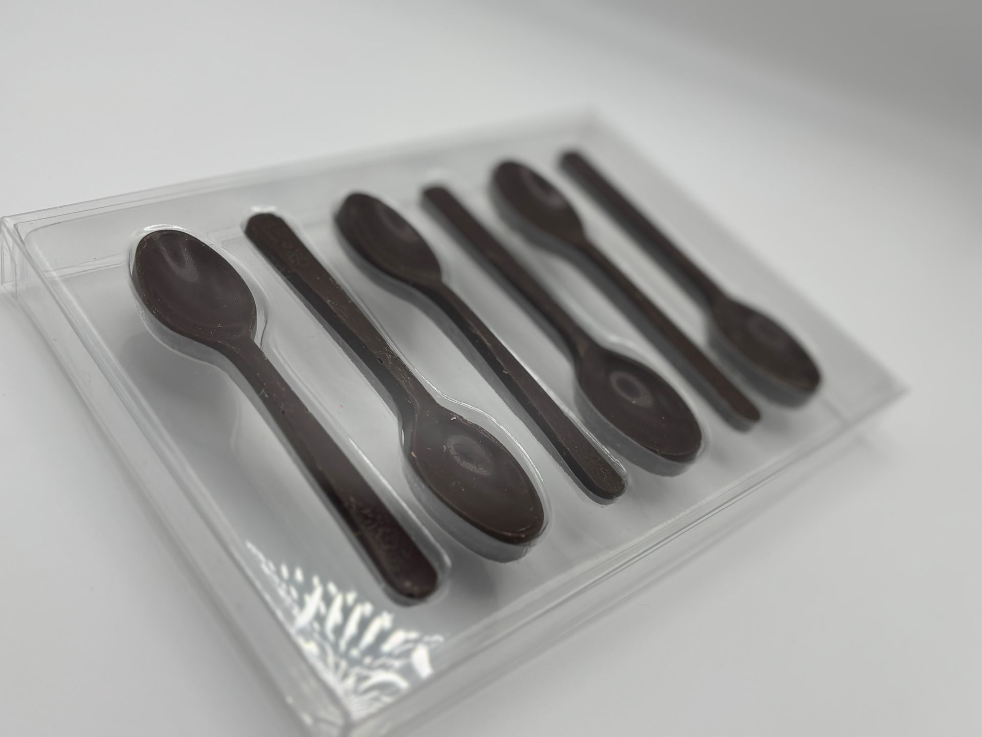 Sei cucchiaini di cioccolato fondente in una confezione di plastica trasparente, disposti in diagonale su uno sfondo bianco.
