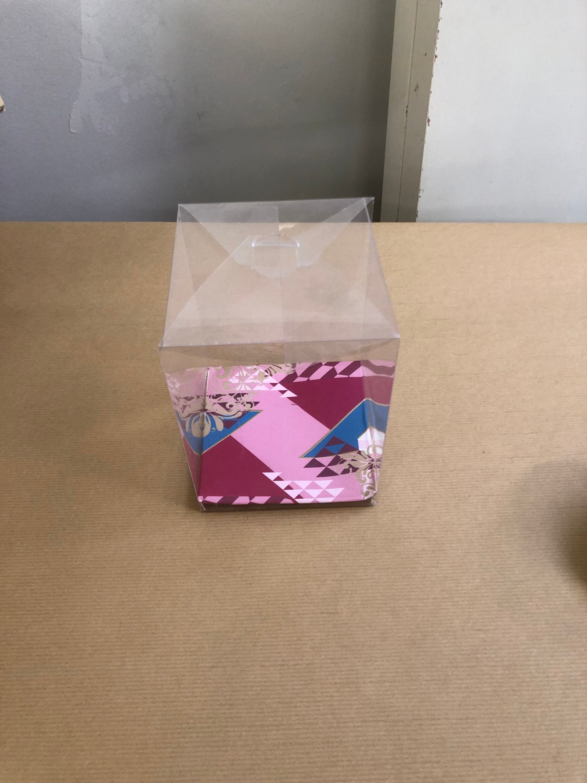 Scatola trasparente con decorazioni rosa, rosse e blu, appoggiata su un tavolo di legno.