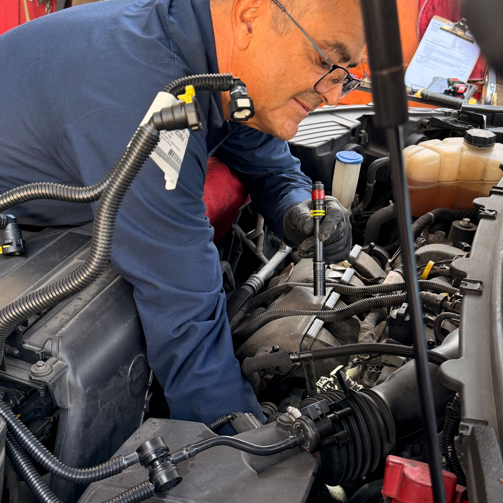 A technician in a navy shirt inspects the engine of a car, holding a tool over the internal components.