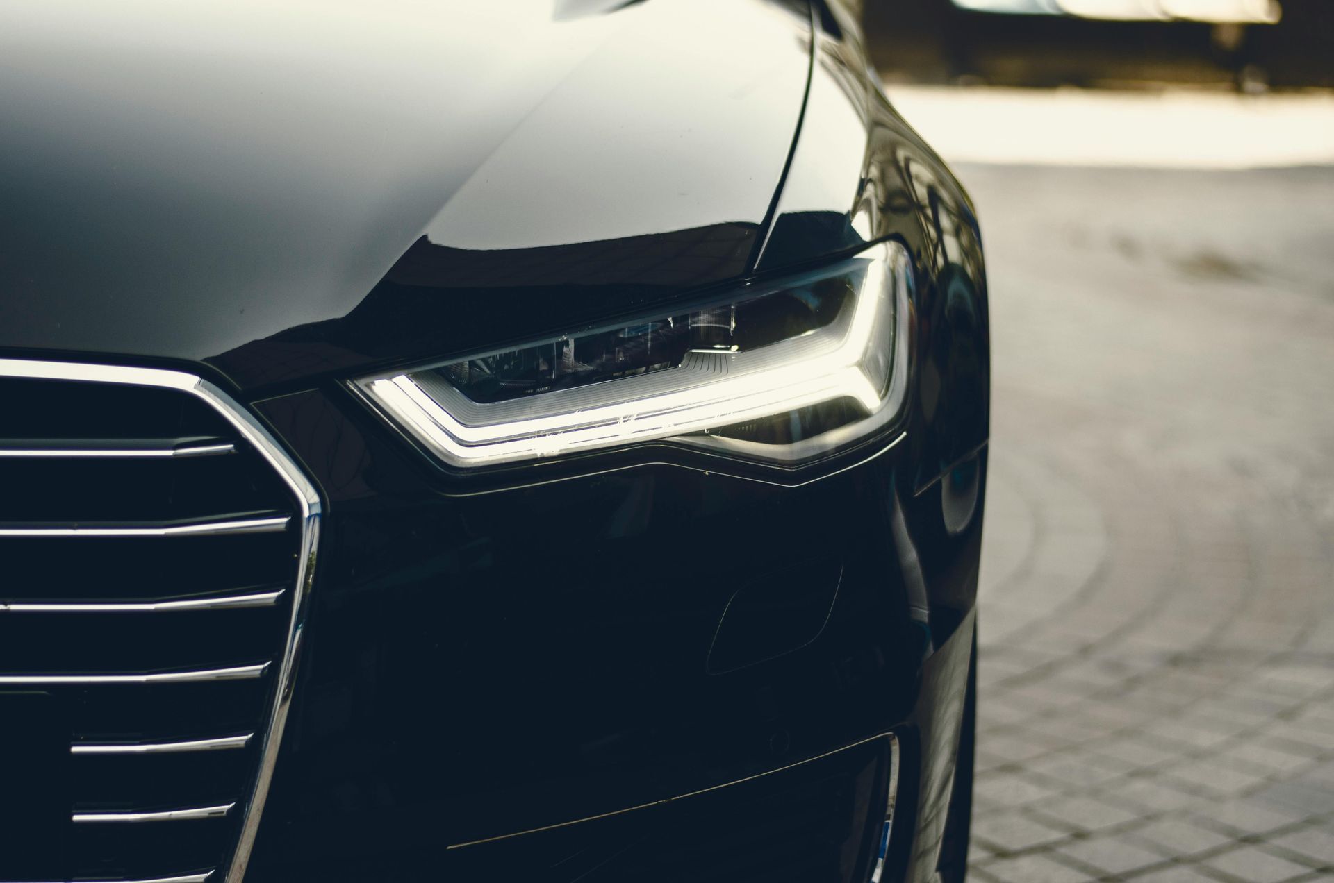 A close-up view of the front headlight and chrome grille of a black Audi parked on a paved surface.