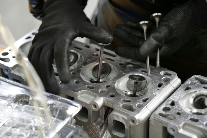 A worker wearing black gloves installs metal engine valves into a cylinder head.