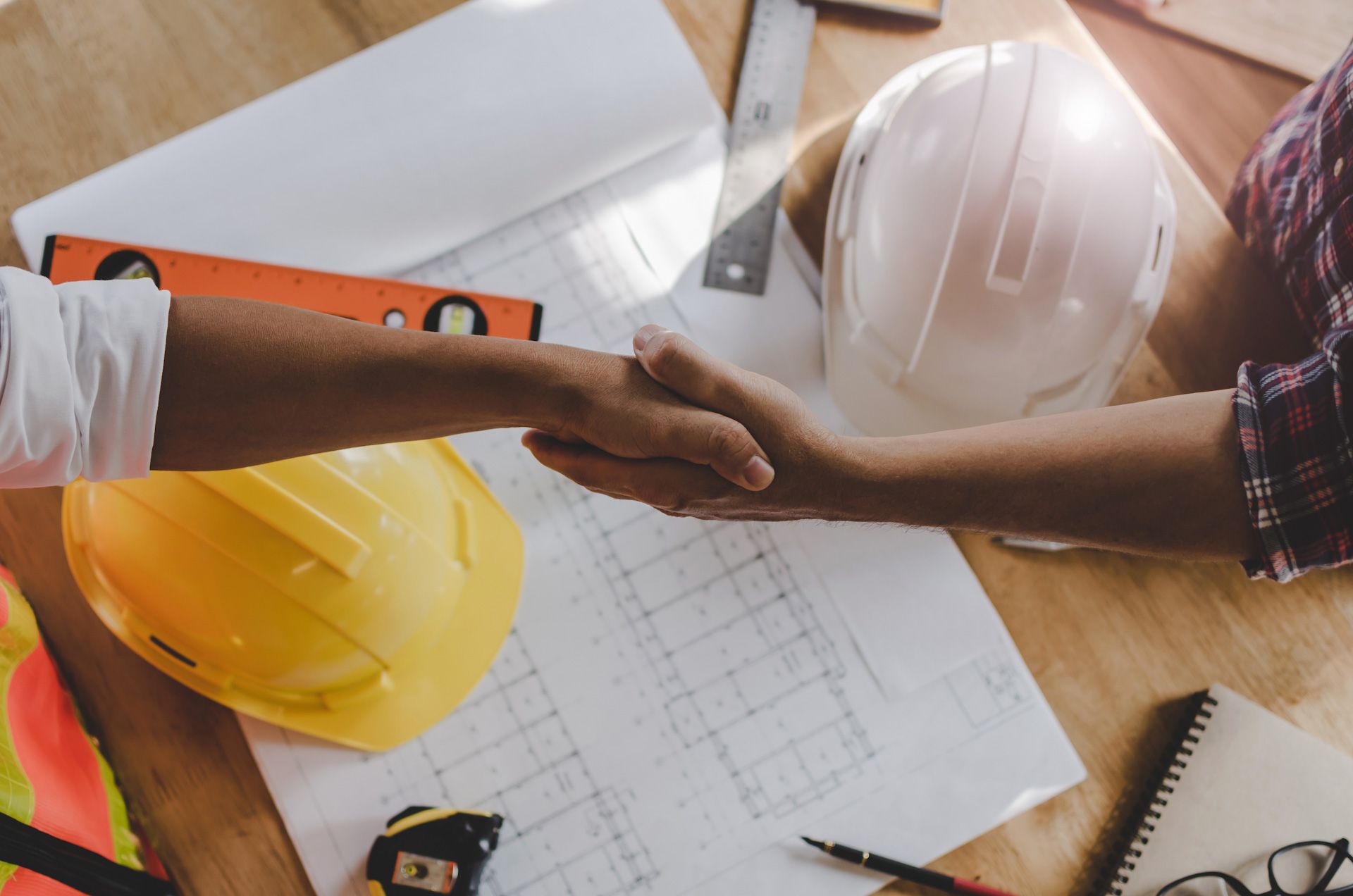 A couple of people shaking hands over a table with hard hats.