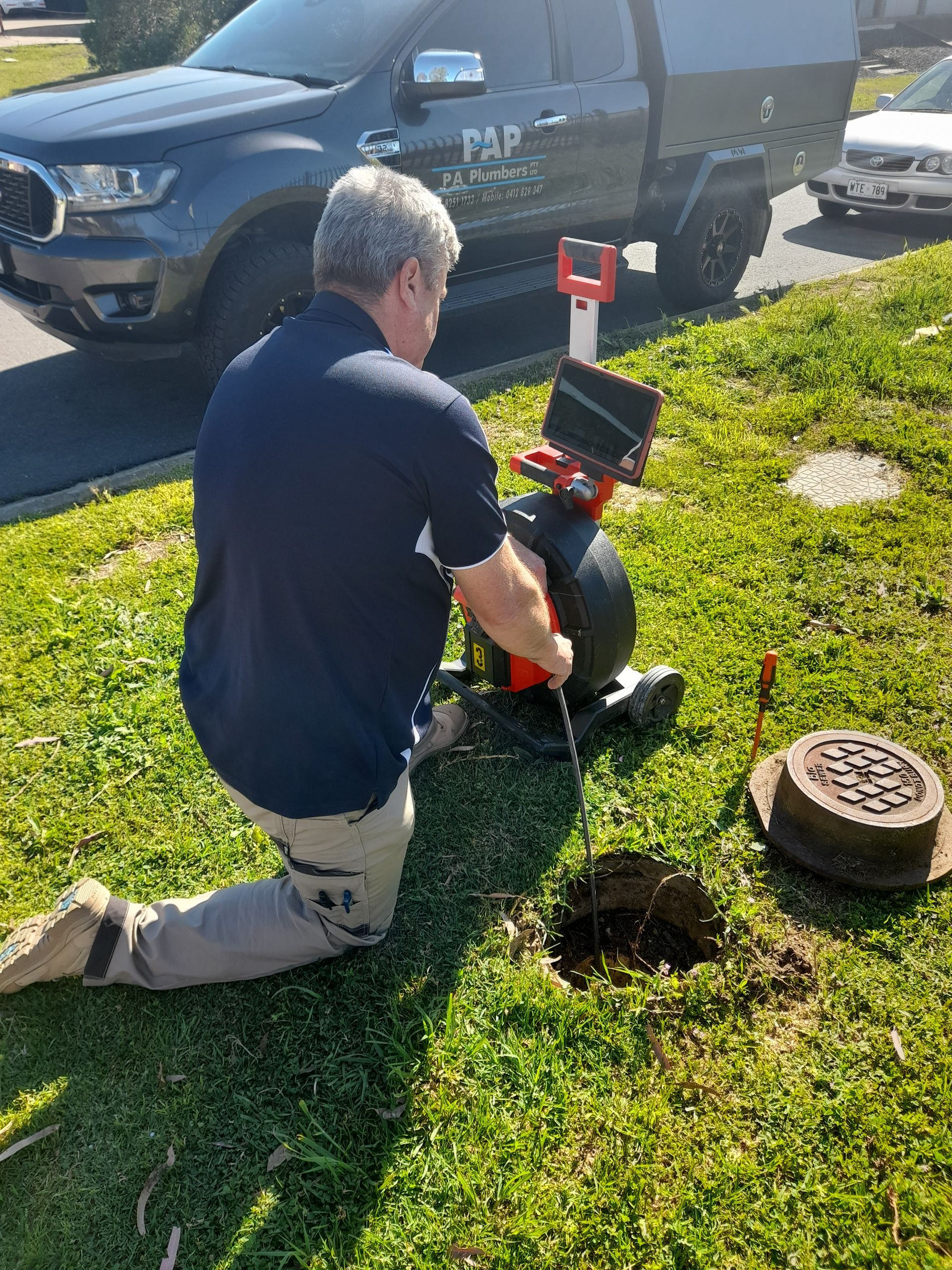 A man is kneeling down in the grass while using a drain camera.