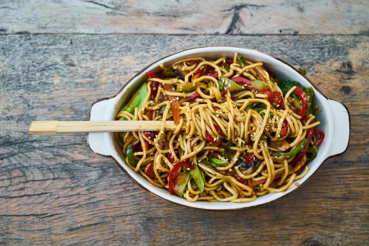 A bowl of noodles with vegetables and chopsticks on a wooden table.