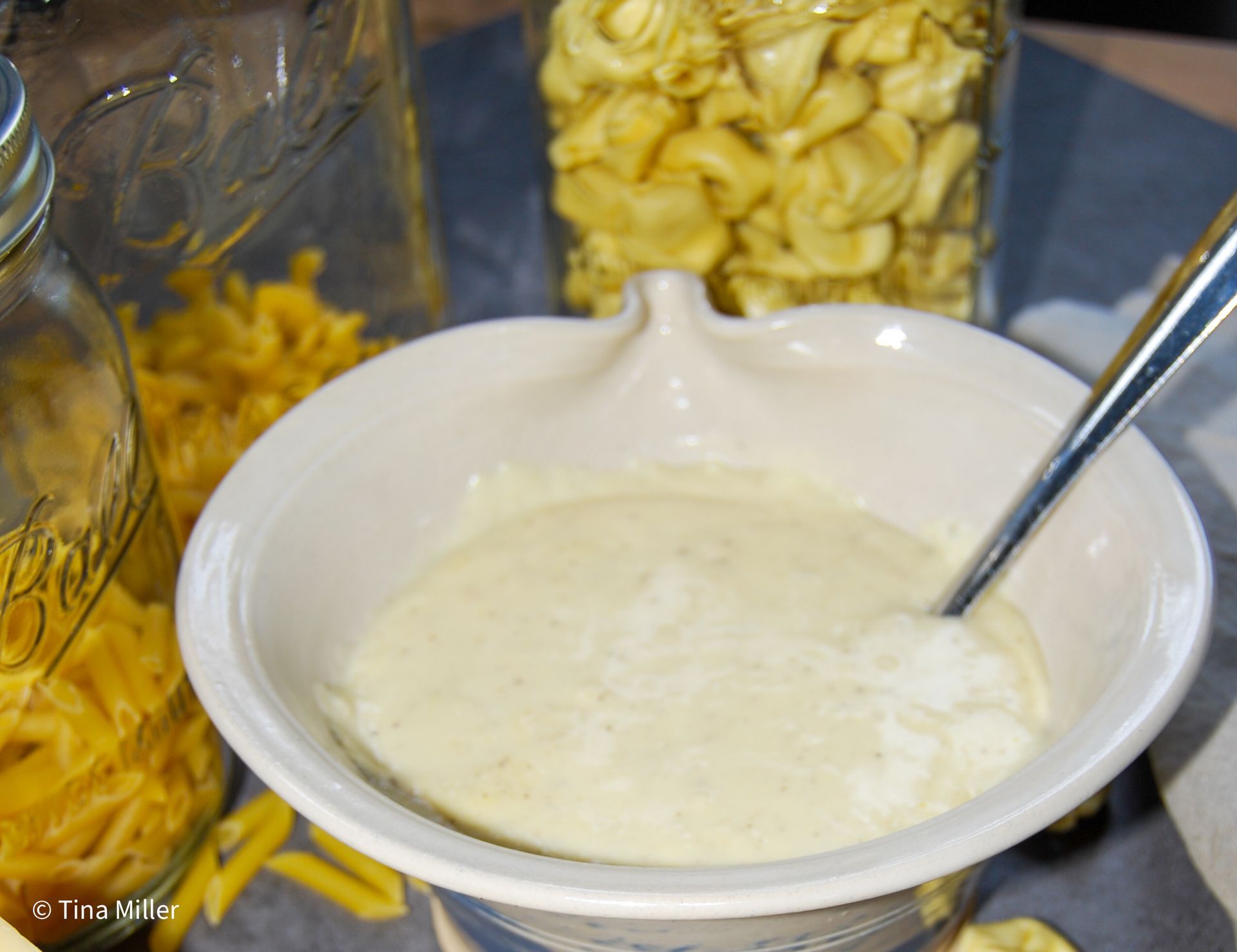 A white ceramic bowl of creamy parmesan Alfredo sauce in front of jars of different types of pasta.