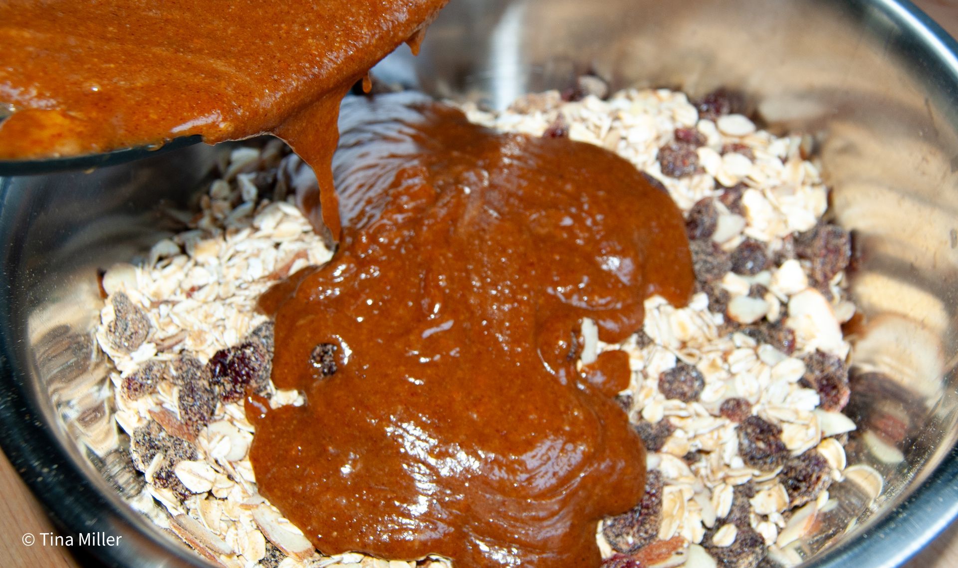 An almond butter glaze being poured over homemade granola mixture with crasinis. 