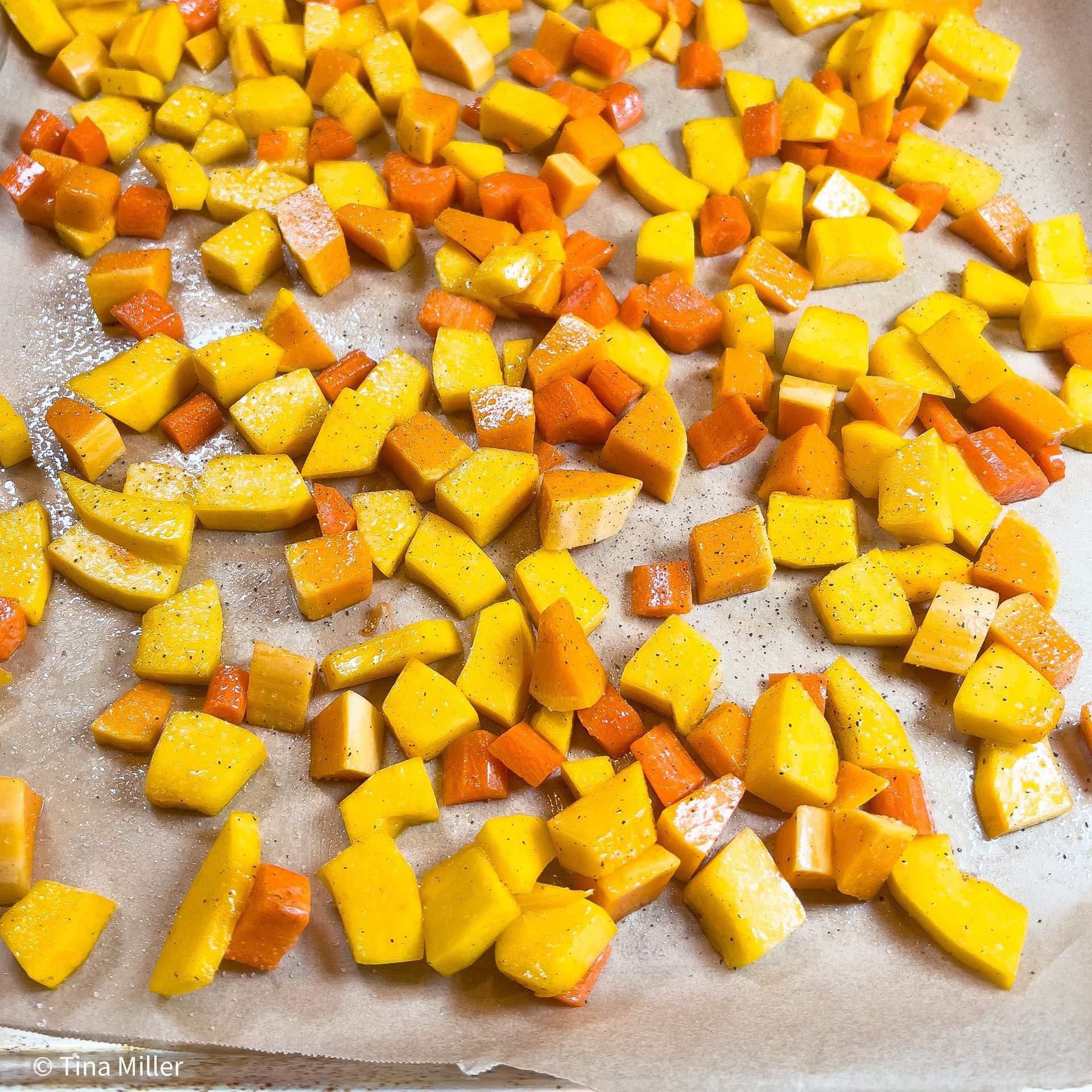 Candy squash, butternut squash, and carrots chopped on a sheet of parchment paper and laying in a single layer on a sheet pan topped with olive oil, salt, and pepper.