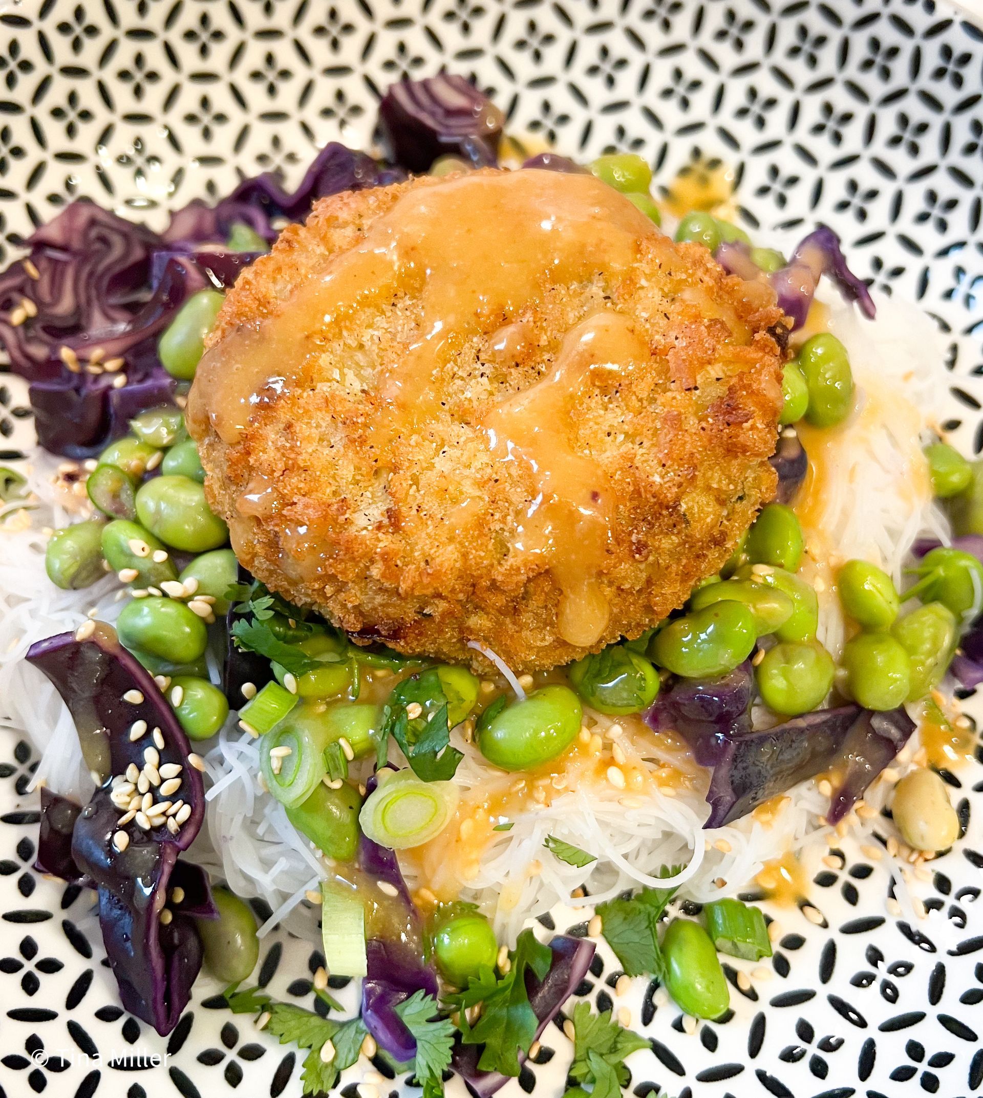 Breaded crab cake on top of red cabbage and edamame slaw with miso dressing served in a black and white bowl.