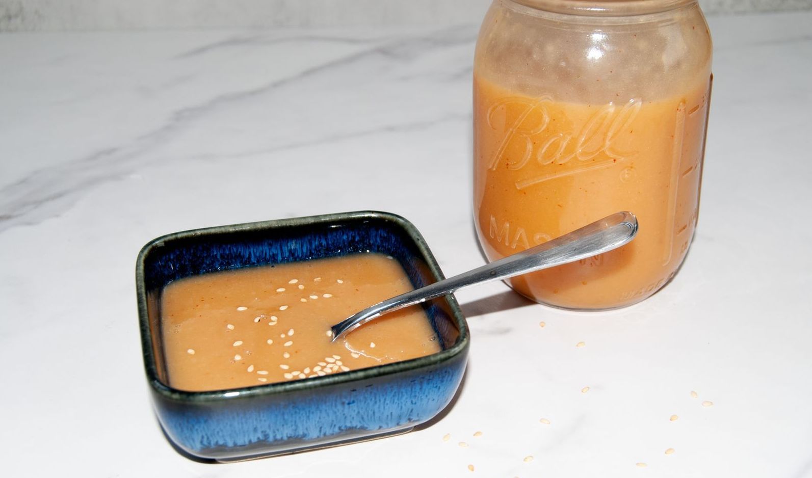 Creamy miso dressing in a small blue bowl with a spoon, garnished with sesame seeds. A jar of dressing in the background.
