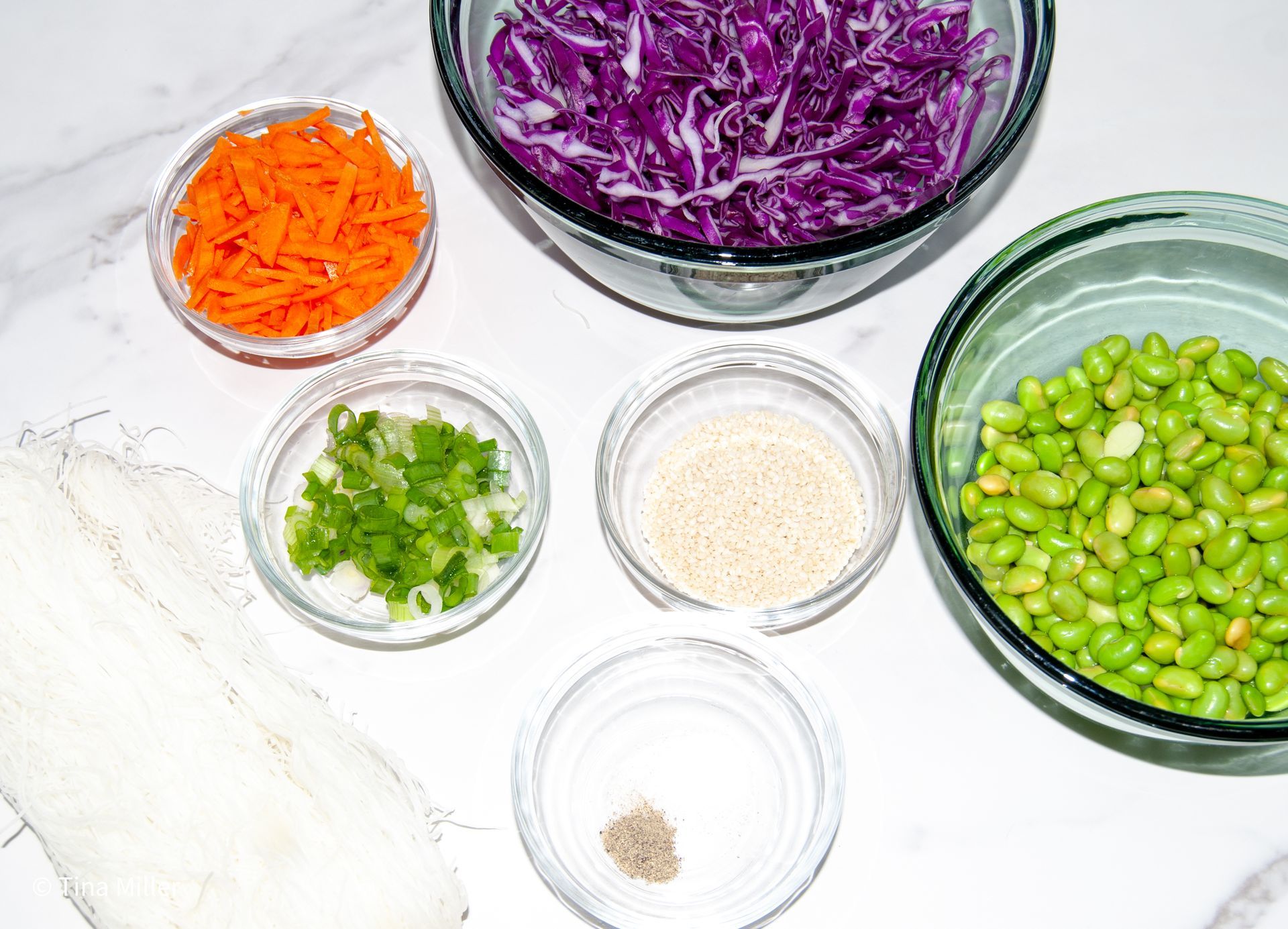 ingredients in glass bowls; Carrot, Red cabbage, Vermicelli on the couter top, Green onion, Sesame seeds, Shelled edamame, Salt and pepper