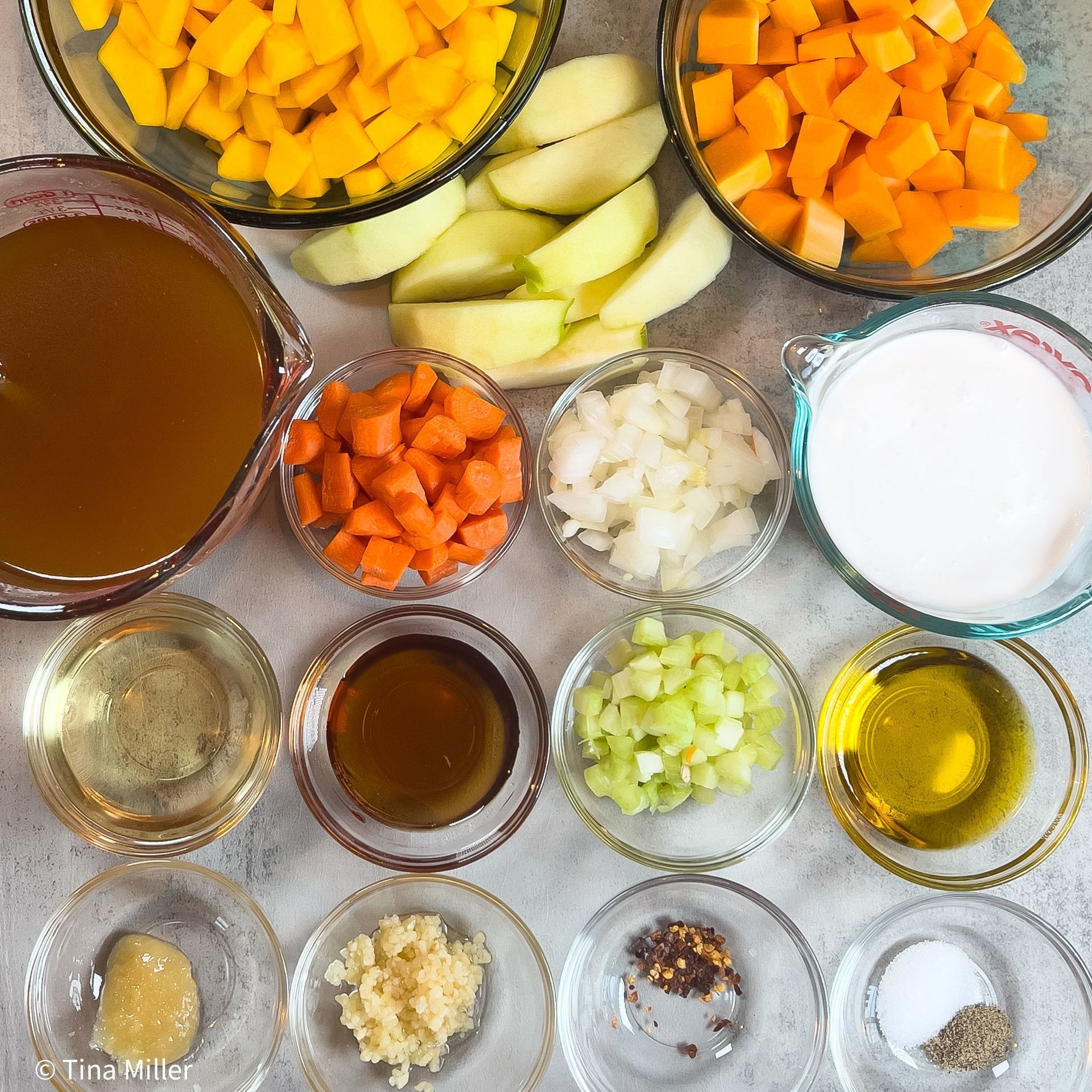 Ingredients for Roasted butternut squash bisque in glass bowls including candy squash, granny smith apples, butternut squash, vegetable broth, carrots, onion, coconut milk, white wine, agave sweetener, celery, olive oil, garlic, ginger, red pepper flakes, salt and pepper.