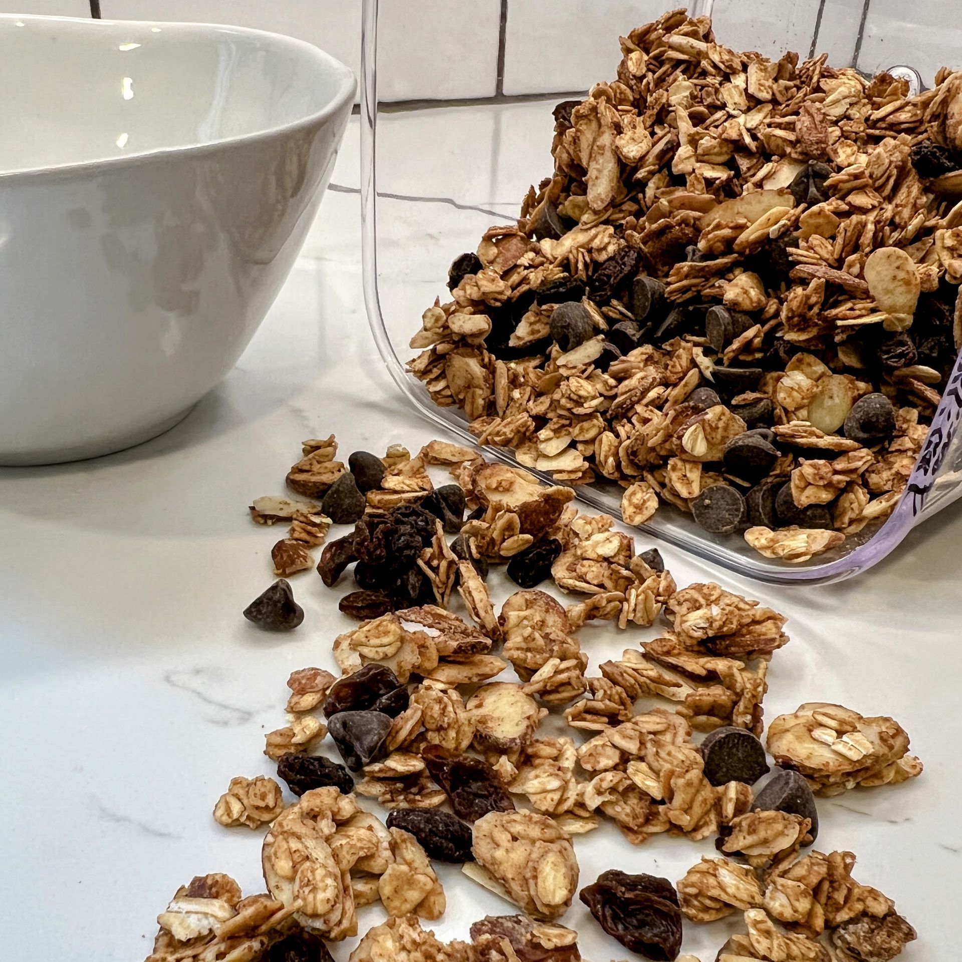 Homemade granola scattered across a white kitchen counter top with a white bowl in the background. 