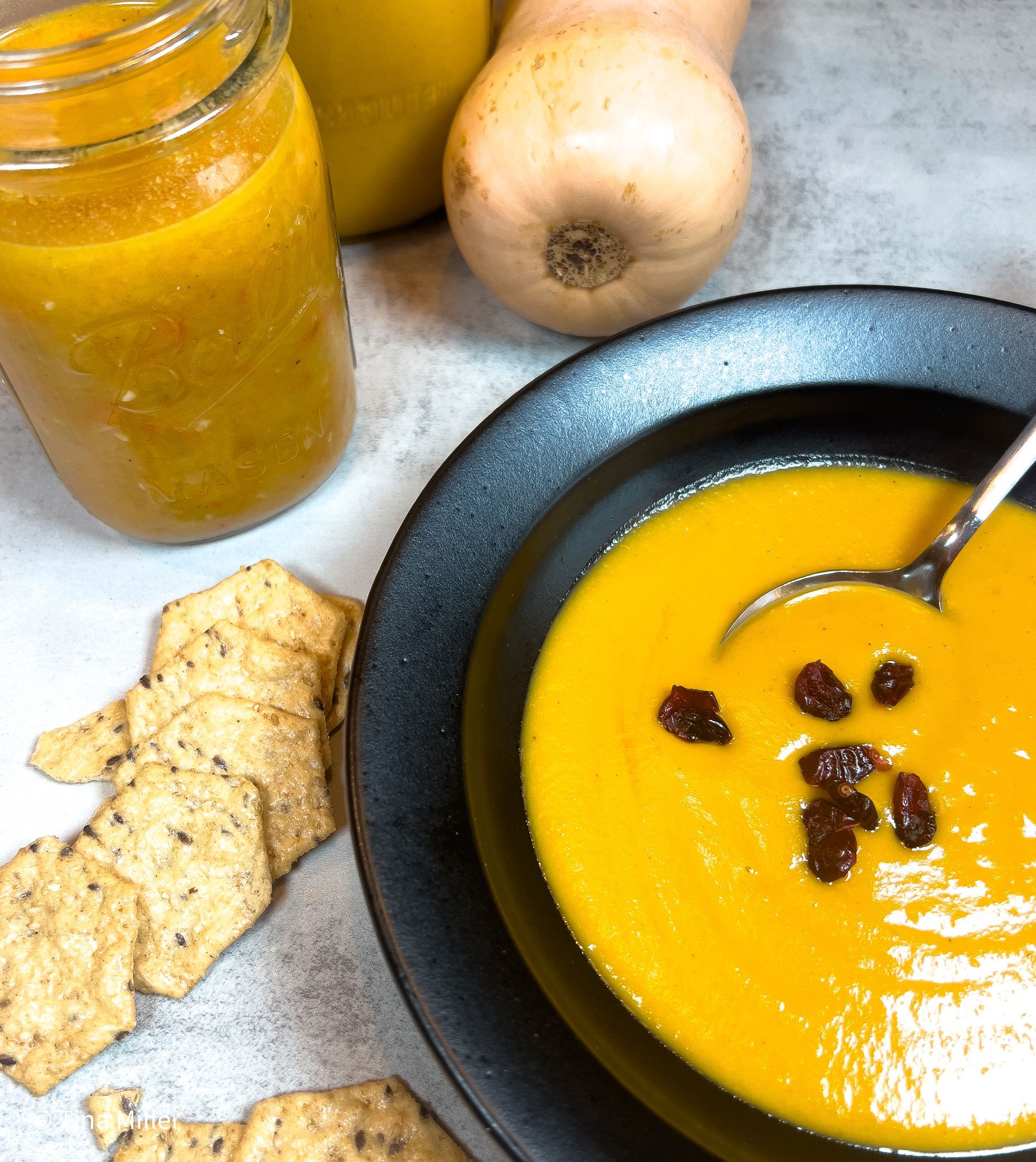 Butternut squash bisque in a black bowl with a spoon, garnished with plump crasins. Crackers and two jars of bisque along with a butternut squash all sitting on the surface by the bowl.