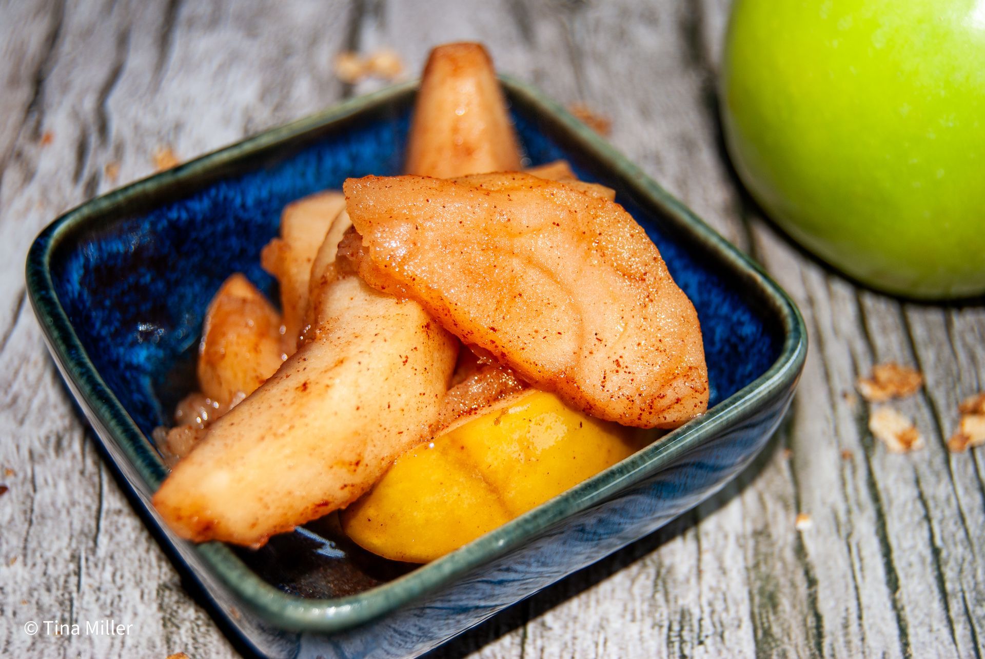 Slices of cinnamon apples in a small blue ceramic bowl on a wooden table with a green apple.