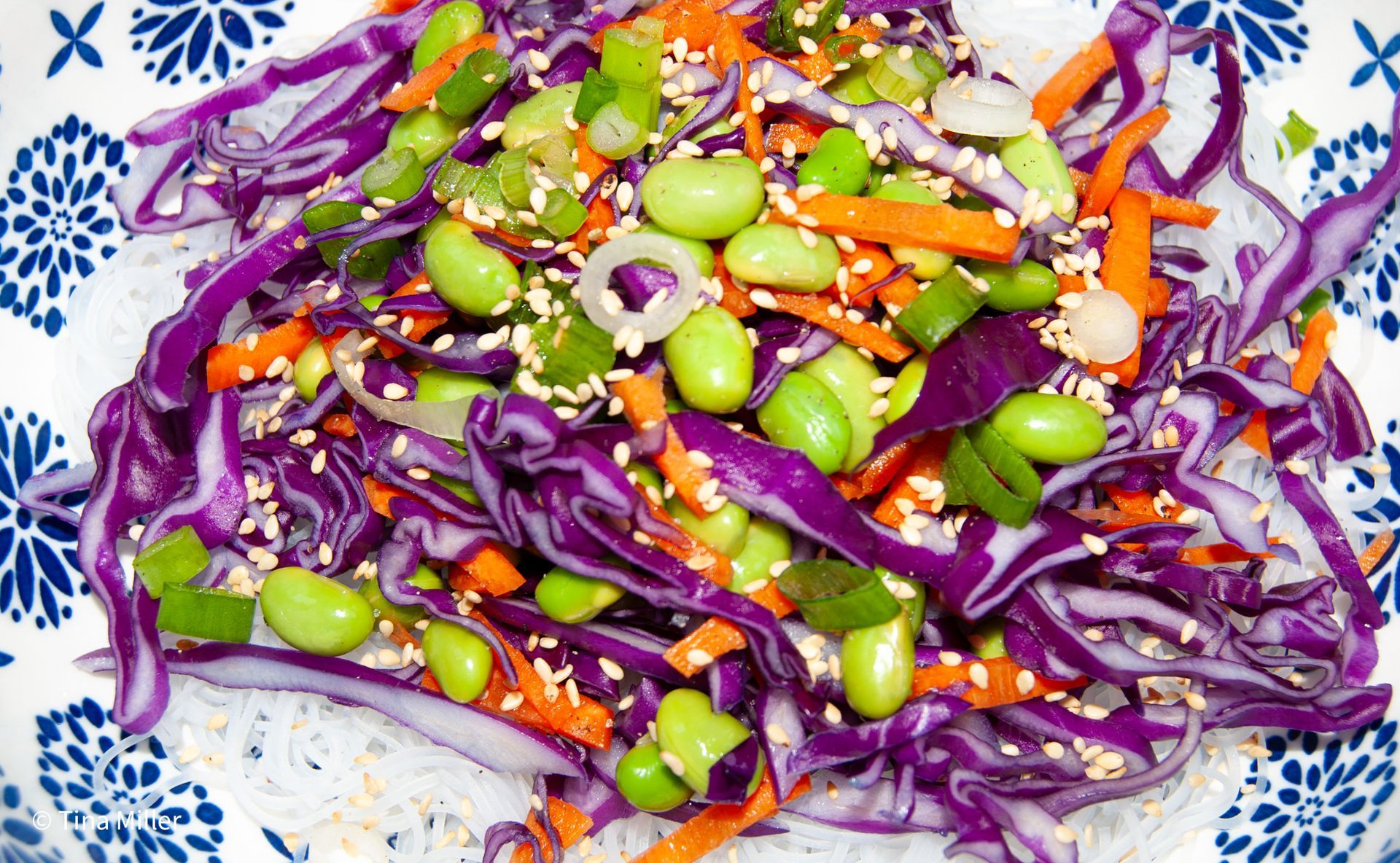 Red cabbage and edamame slaw with carrots in a bowl over rice vermicelli, garnished with sesame seeds and miso dressing.