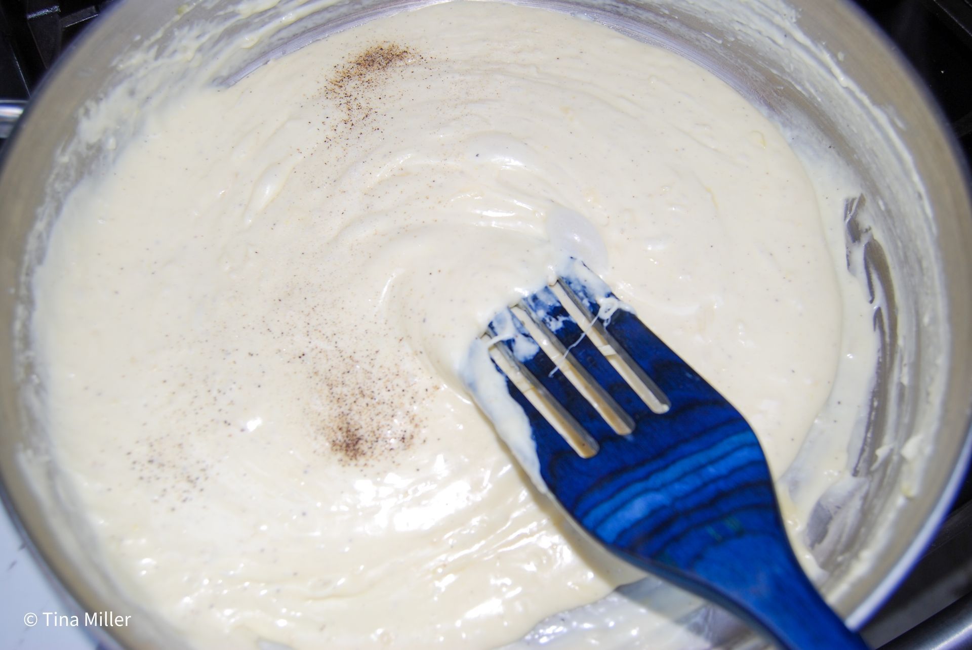 A creamy cheese sauce mixture with added pepper being stirred with a blue wooden spatula.