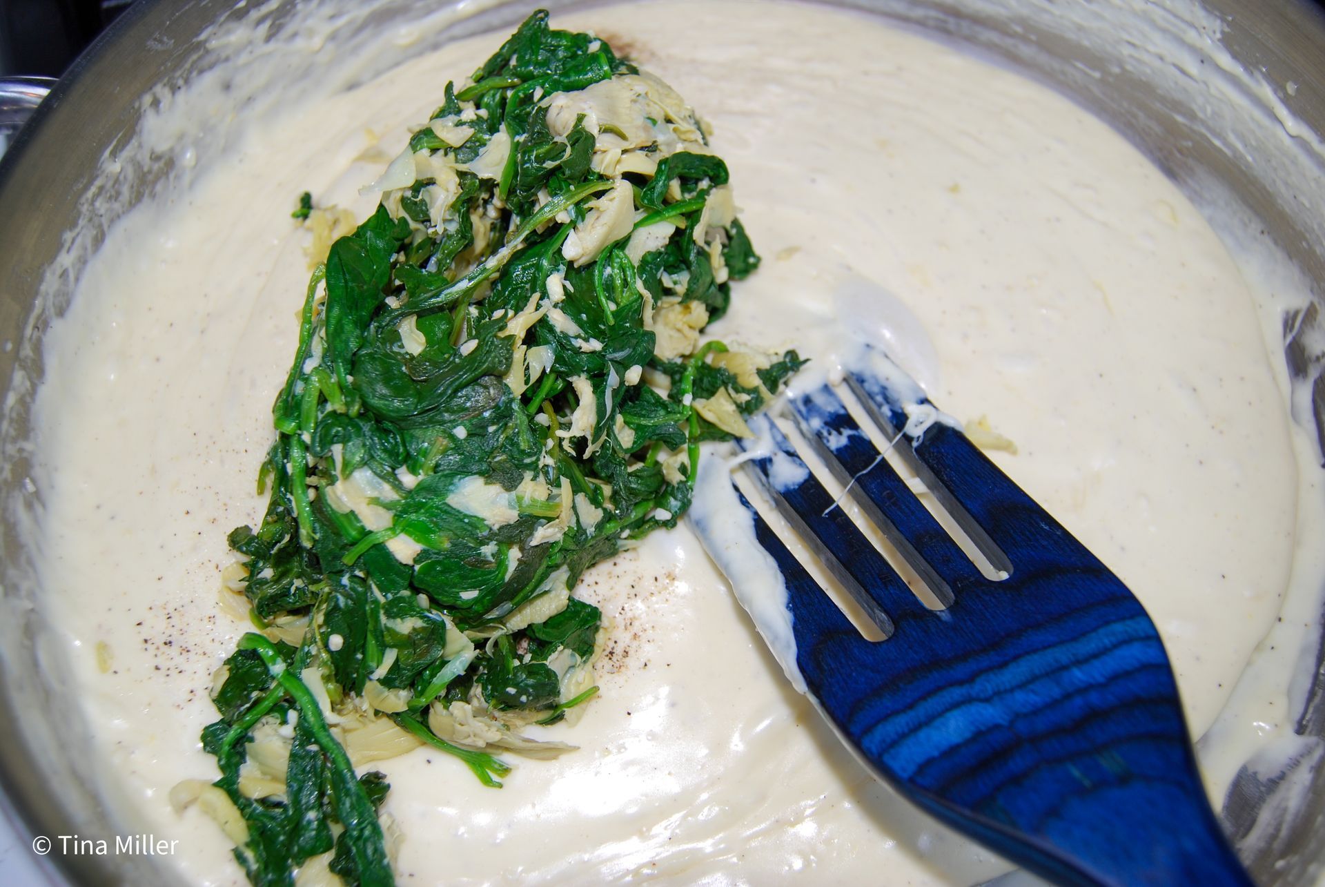 sautéed spinach and artichoke hearts being added to a creamy cheese mixture and being stirred with a blue wooden spatula.
