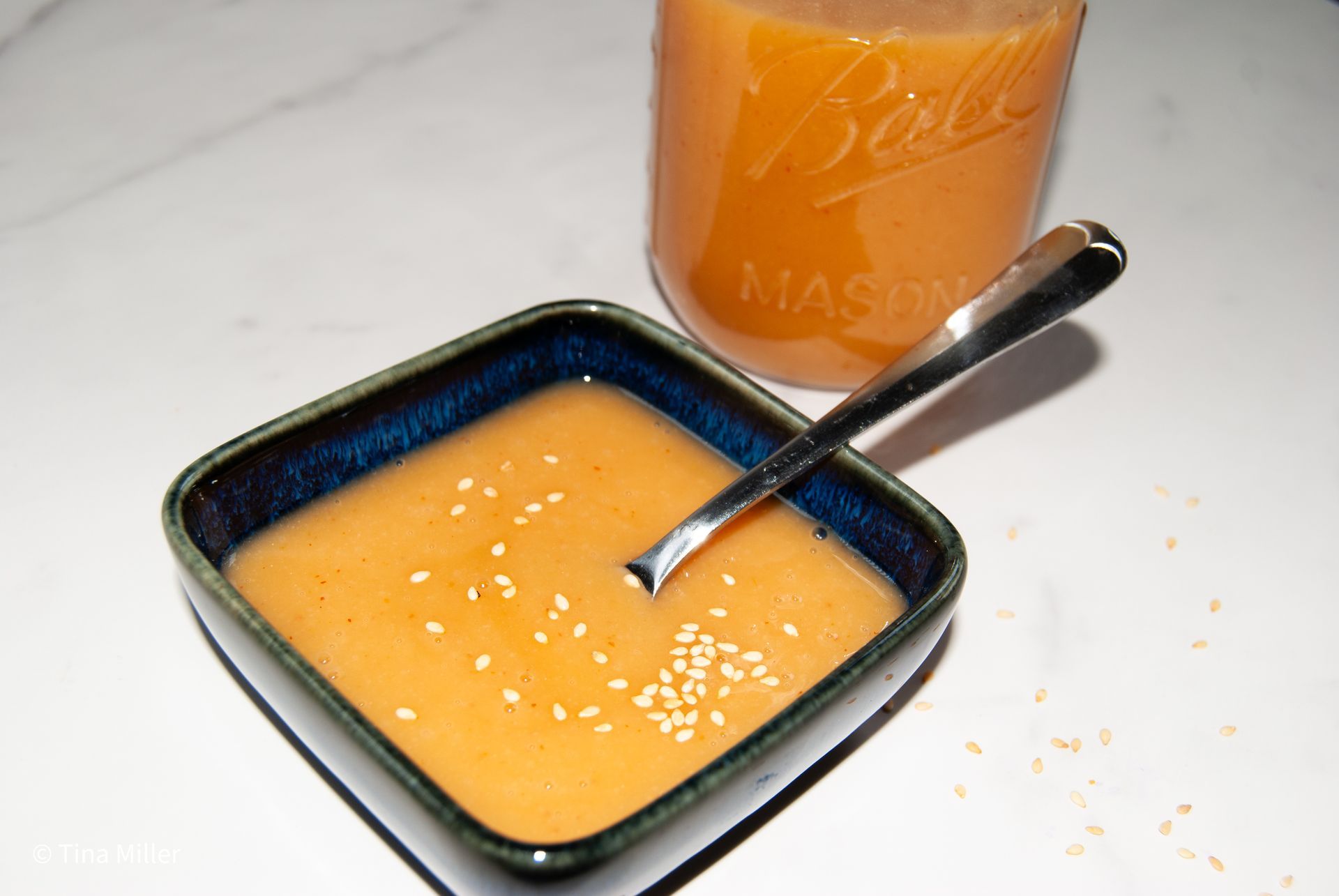Creamy Asian inspired miso dressing in a blue ceramic bowl with a spoon for dipping, garnished with sesame seeds. A jar of dressing in the background.