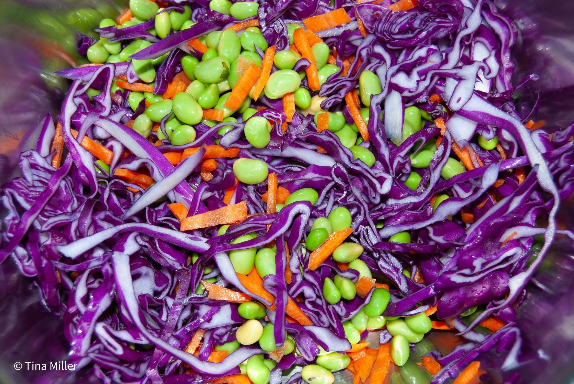 Bright green shelled edamame, shredded red cabbage, and orange julienne carrots tossed together in a metal mixing bowl.