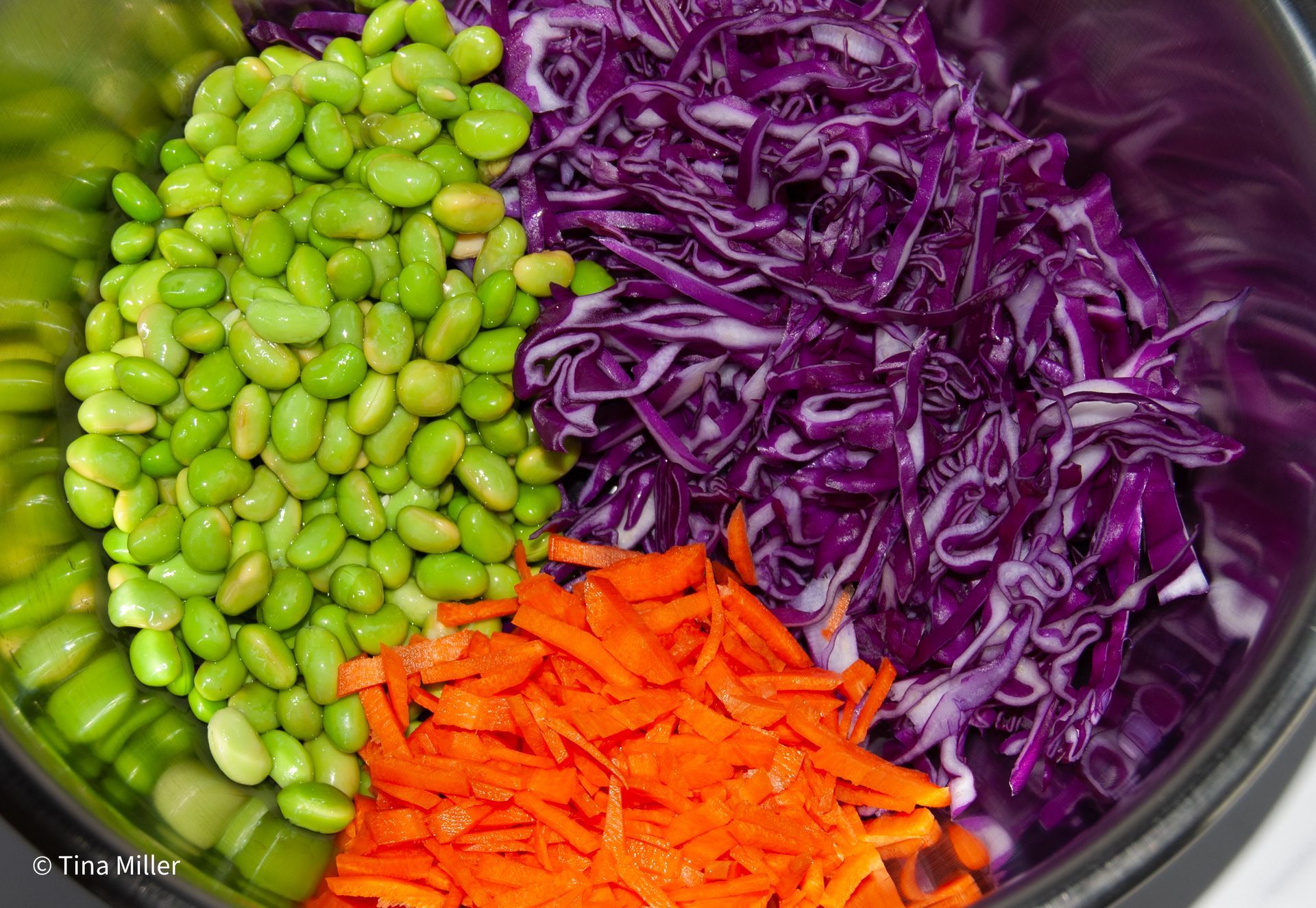 Bright green shelled edamame, shredded red cabbage, and orange julienne carrots in a metal mixing bowl.