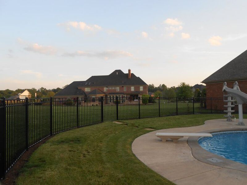 Backyard pool with curved concrete deck, black fence, and house in the background at sunset