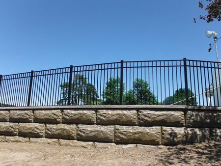 Stone retaining wall with black metal fence against a blue sky and trees