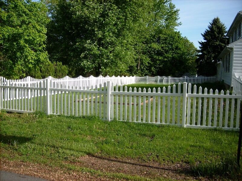 White picket fence lining a grassy yard beside a house and large green trees