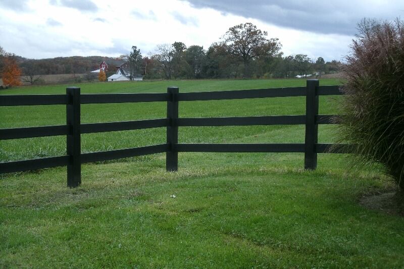 Black wooden fence in a green grassy field with trees and cloudy sky in the background