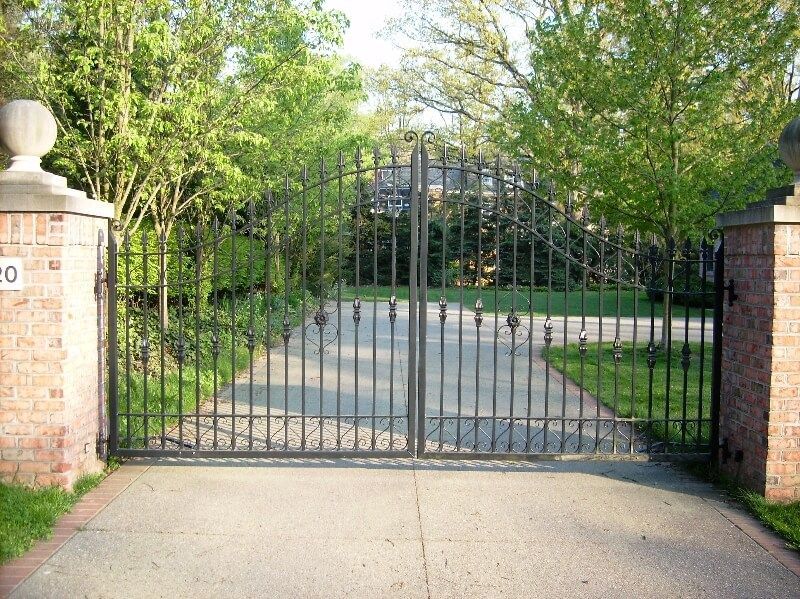 Black iron gate across a driveway entrance between brick pillars and trees